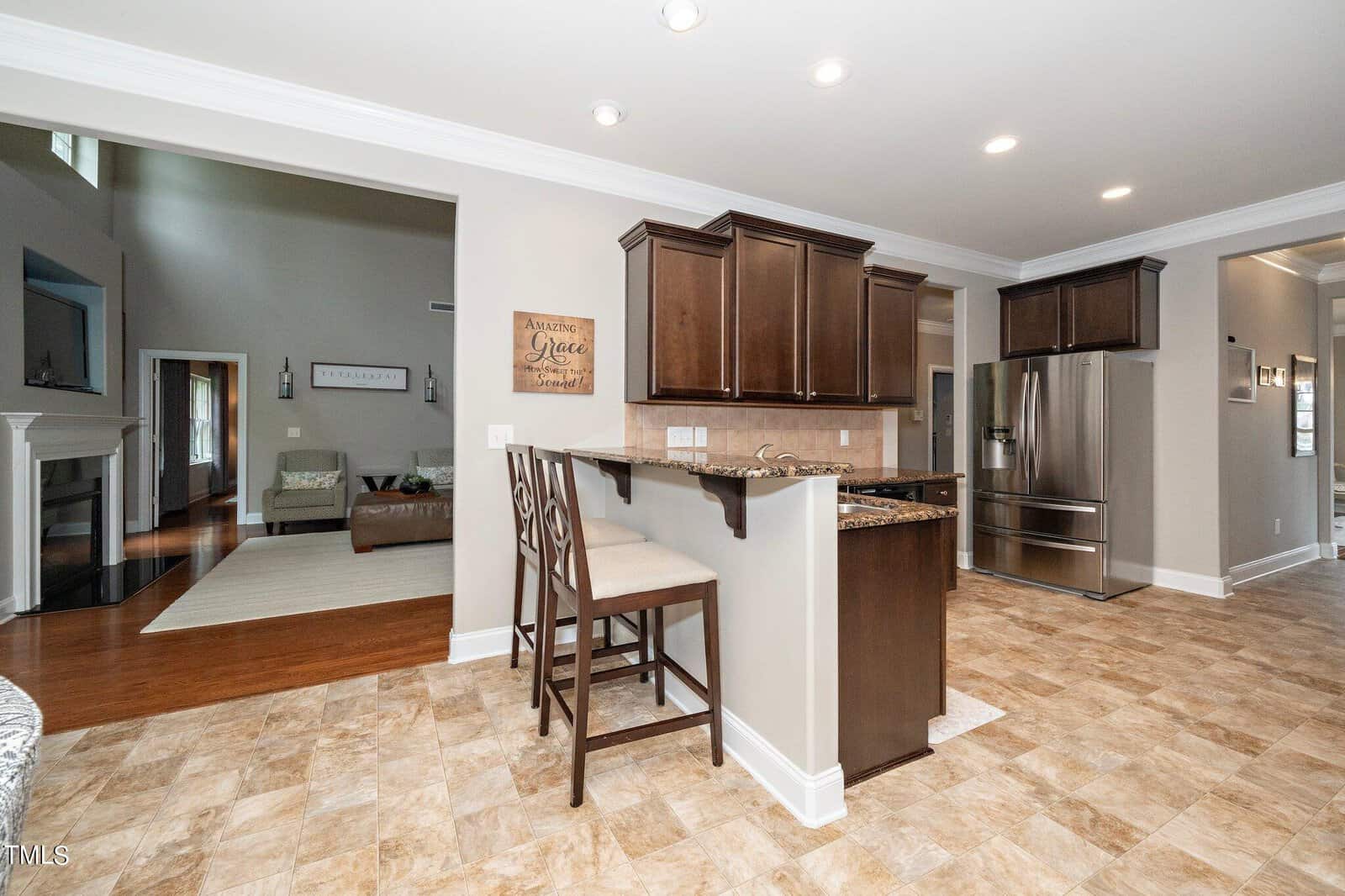A modern kitchen with wooden cabinets and granite countertops. A bar-style seating area with two chairs is in the foreground. Stainless steel appliances, including a refrigerator, are visible. The adjacent living room features a sofa and a fireplace.