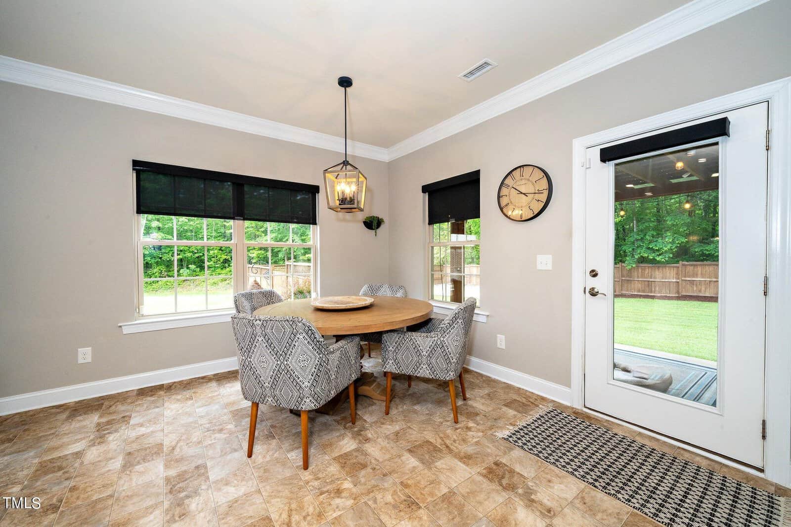 A cozy dining area features a round wooden table with gray upholstered chairs on a tiled floor. Black blinds cover the windows, and a modern lantern-style light hangs overhead. A door with a window leads to a grassy backyard. A clock adorns the wall.