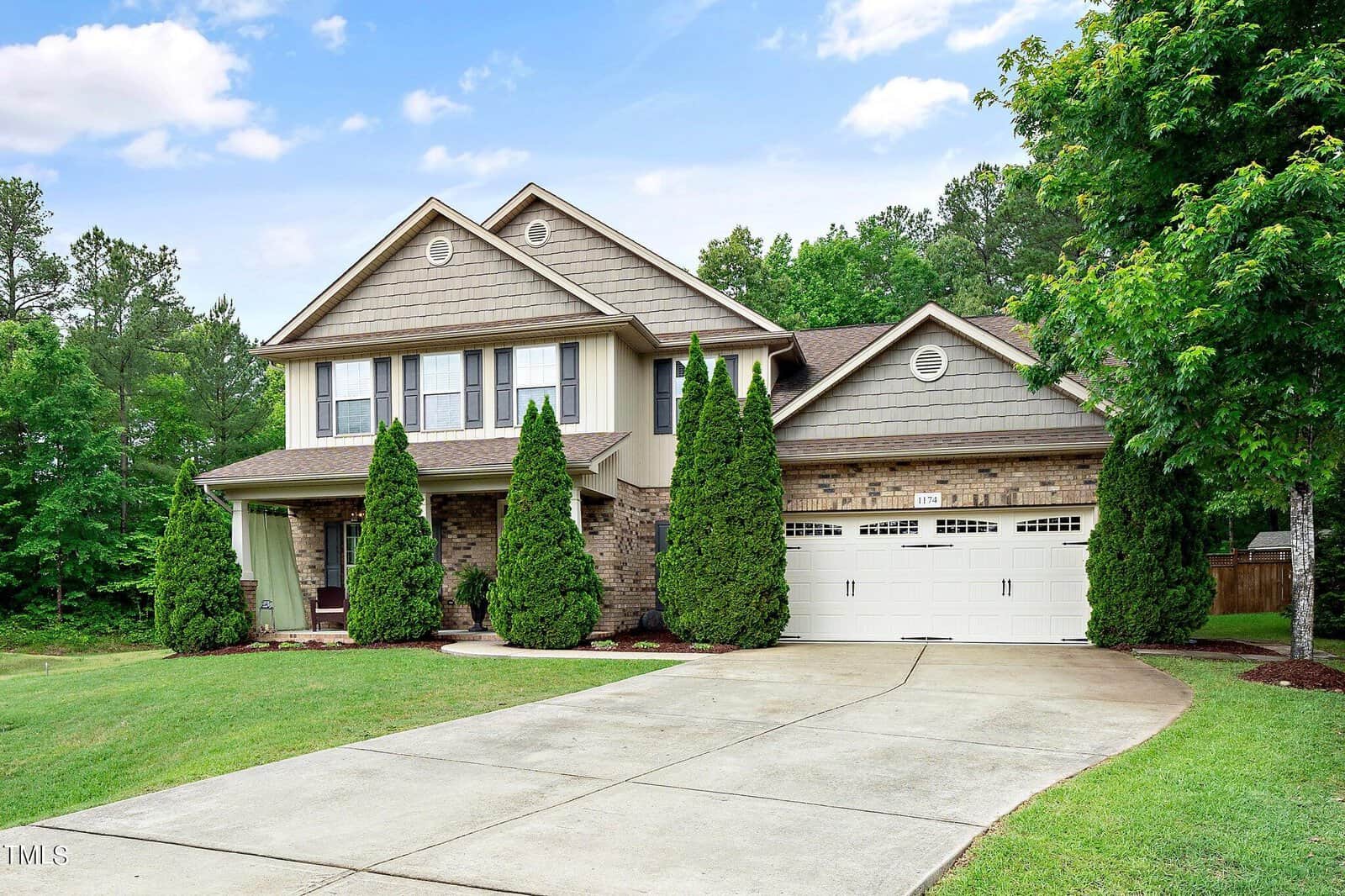 A two-story suburban house on 1174 Jackson Court boasts a brick and siding exterior, double garage, and driveway. The front yard features neatly clipped grass with tall trees near the entrance. Located in Creedmoor, the sky is partly cloudy with trees adorning the background.