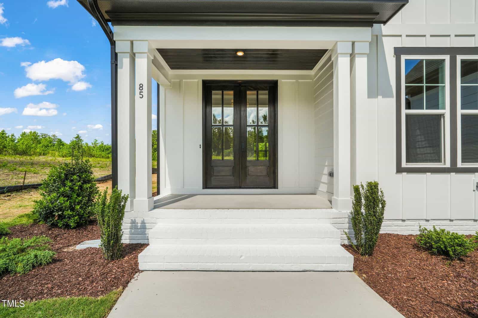 Front porch of a modern white house with double glass doors, white steps, neatly trimmed bushes, and a well-maintained yard on a sunny day.