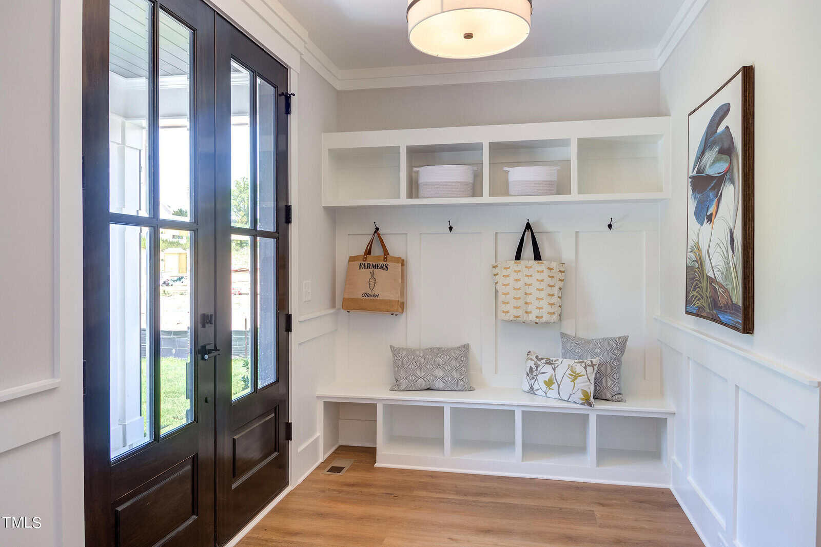 A bright entryway with double glass doors, a built-in white bench with cubbies, pillows, storage baskets, and hooks holding bags. A large bird artwork hangs on the wall above wainscoting.