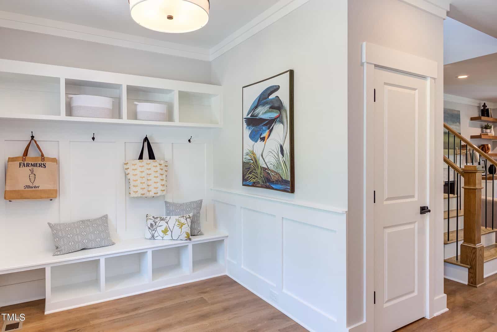 A bright, modern mudroom with white built-in cubbies, benches with gray and floral pillows, baskets on top, and two hanging bags. A large bird artwork decorates the wall above wainscoting. A staircase is visible to the right.