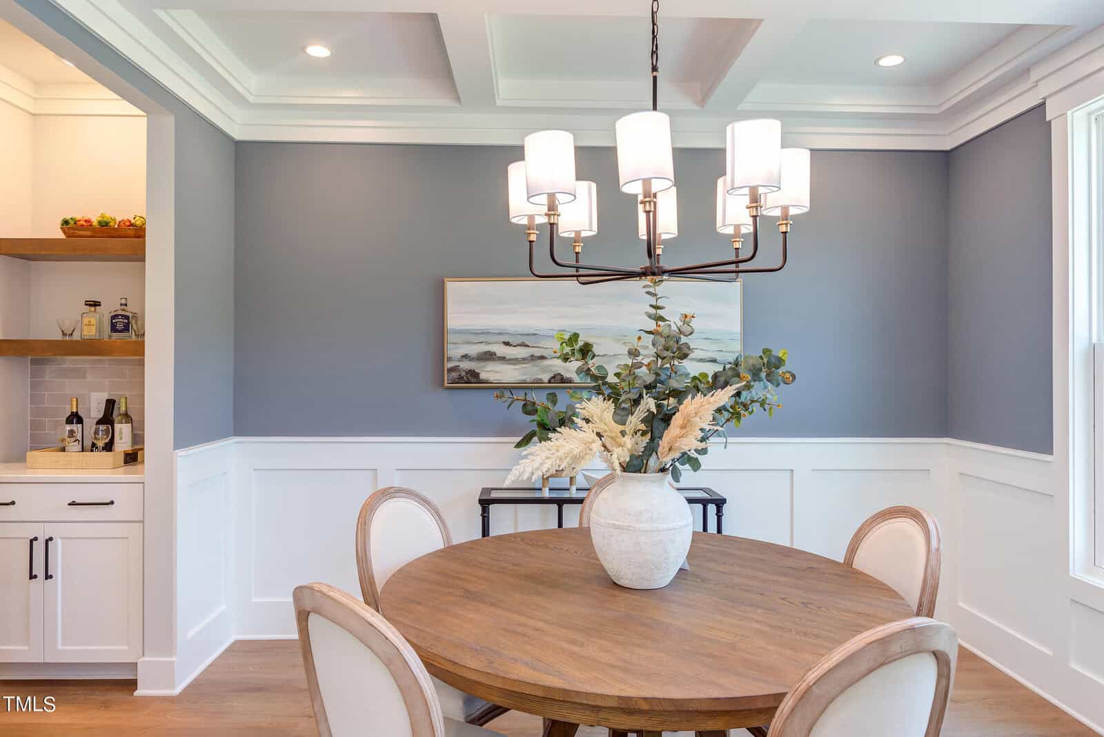 A modern dining room with a round wooden table, four upholstered chairs, a vase with greenery as a centerpiece, a blue accent wall with white wainscoting, and a chandelier above. A sideboard and wall art are in the background.