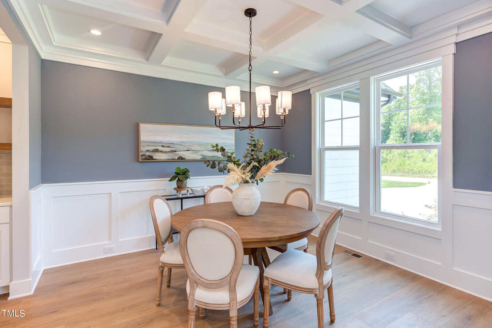 A bright dining area with a round wooden table, six upholstered chairs, a large vase with dried flowers, modern chandelier, gray-blue walls with white wainscoting, a window, and a landscape painting.