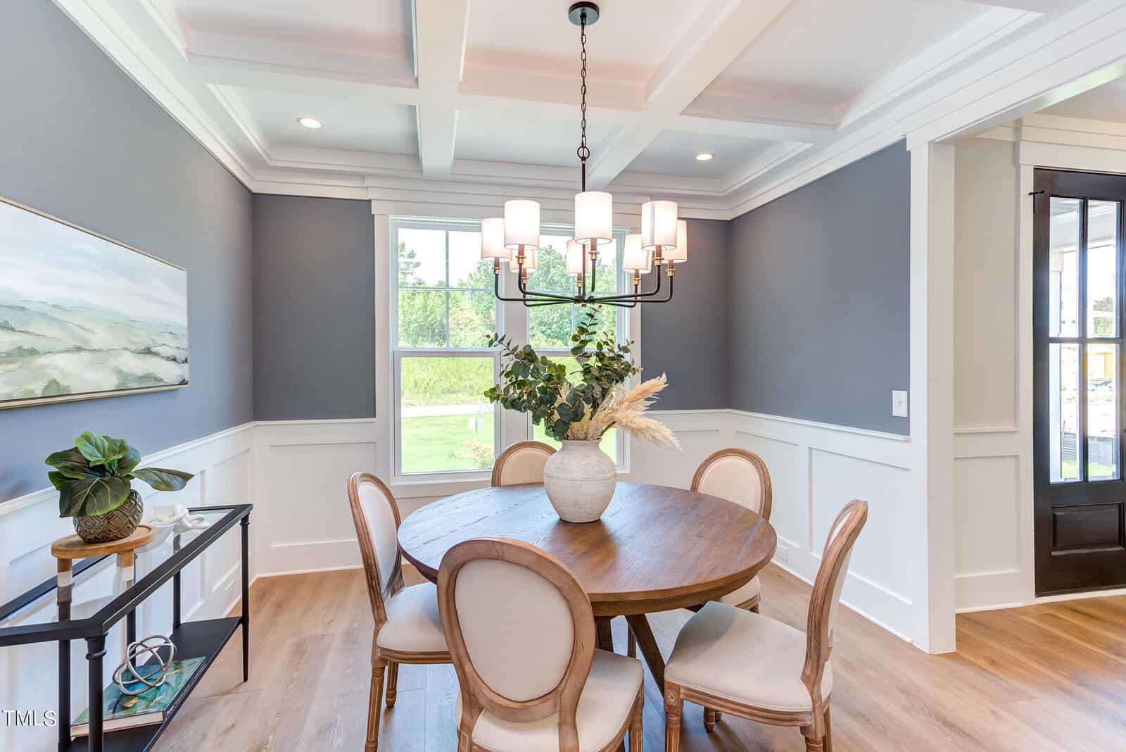 A dining room with gray walls, white wainscoting, a round wooden table with four chairs, a chandelier, and a large window letting in natural light. A vase with greenery is on the table; art and decor are on the side table.