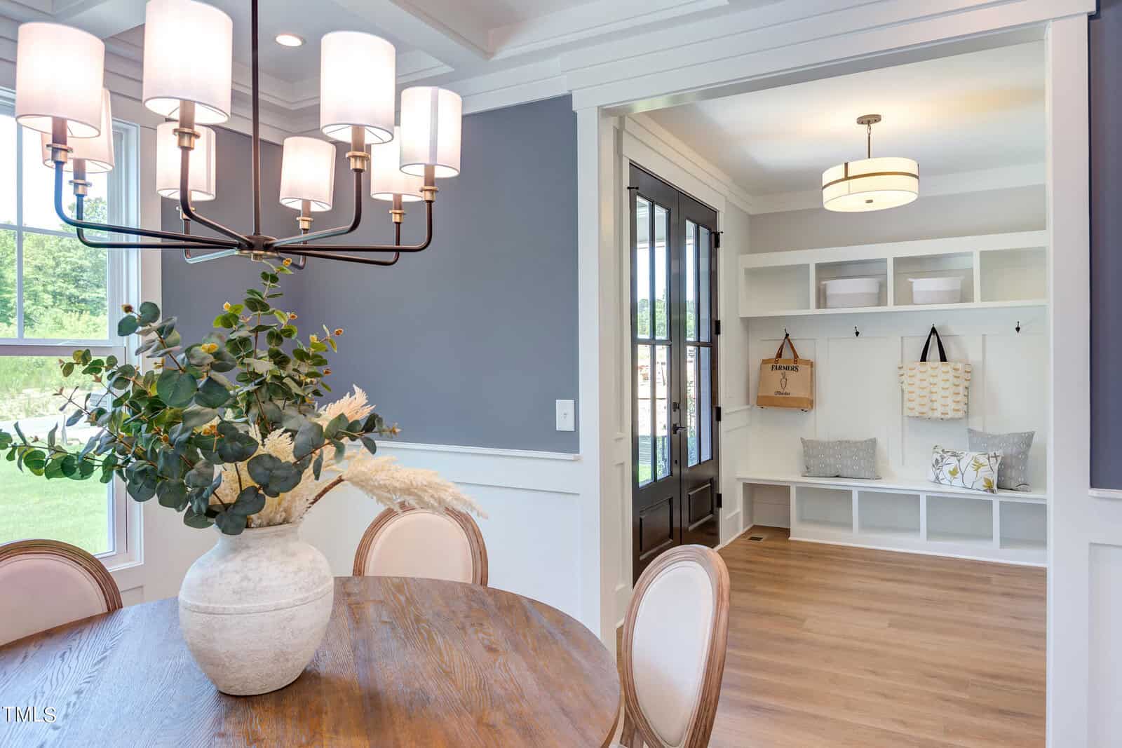 A bright dining area with a round wooden table, cushioned chairs, a modern chandelier, and a vase with greenery. In the background, a mudroom with cubbies, hooks, tote bags, and pillows is visible.
