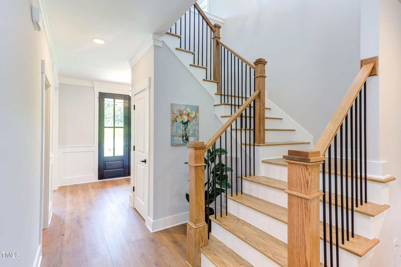 Bright entryway with wooden floors, a staircase with wooden railings and black spindles, a black front door with glass panels, white walls, a painting on the wall, and a plant by the stairs.