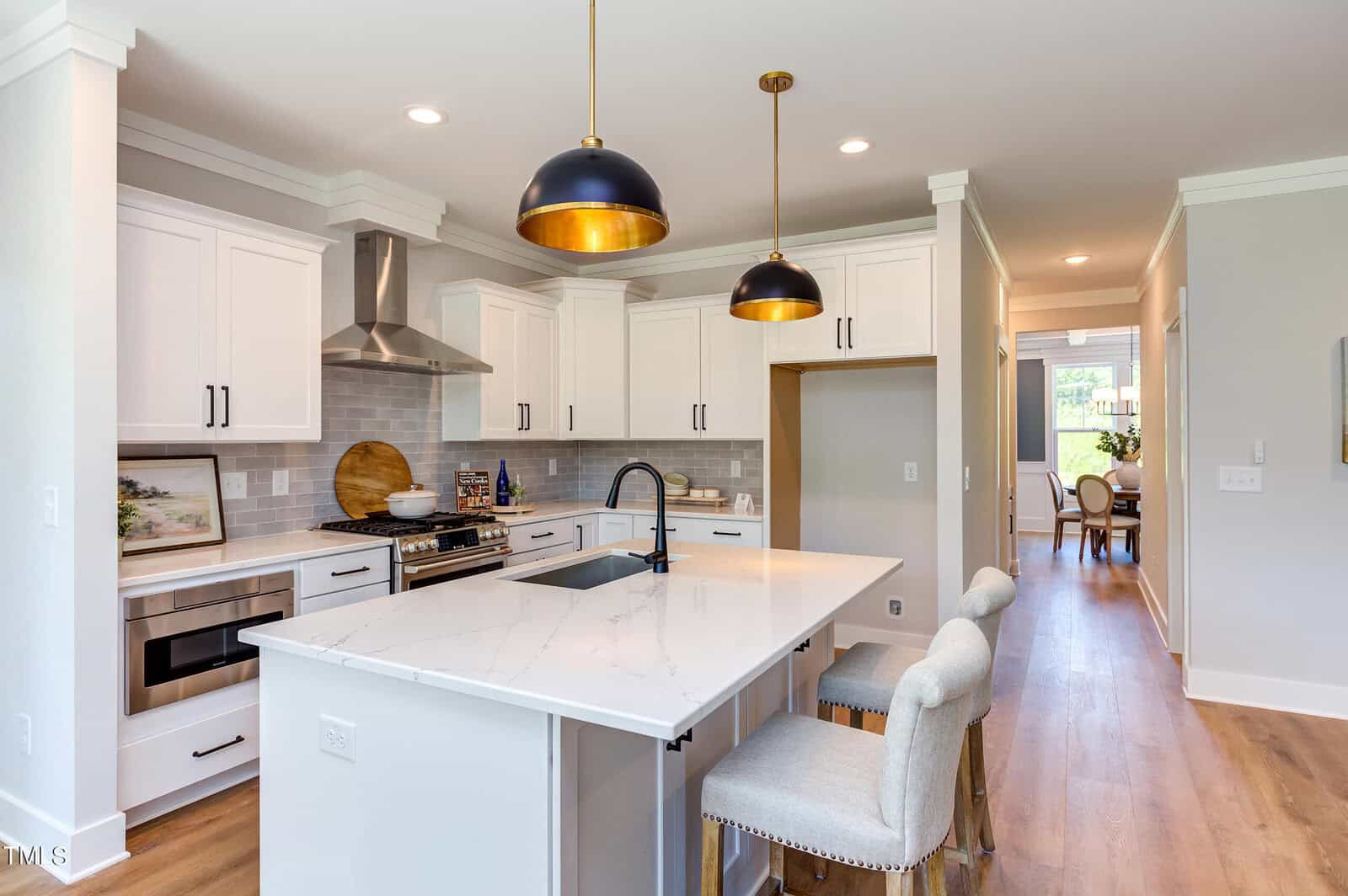 Modern kitchen with white cabinets, marble island, and black fixtures. Two cushioned stools sit at the island. Gold and black pendant lights hang above. Stainless steel appliances and wood floors complete the space.