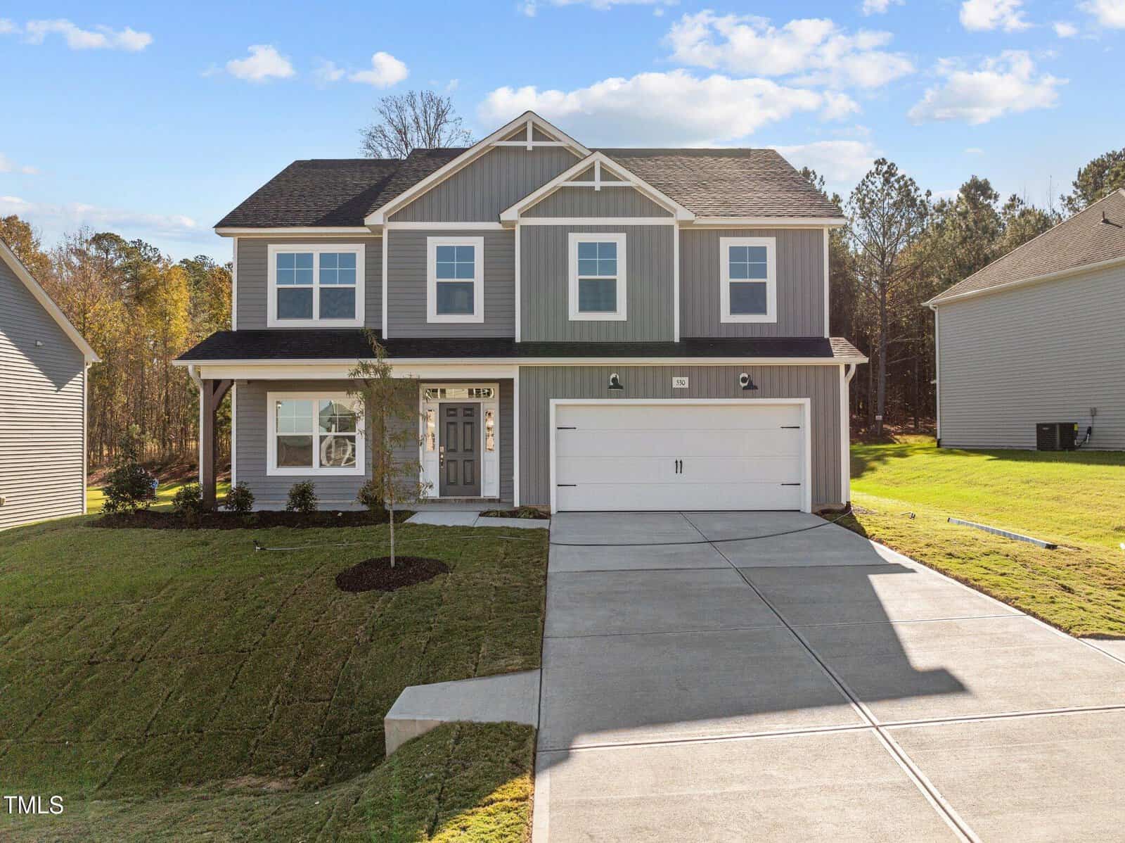 A two-story modern gray house with white trim, a covered front porch, double garage, and a neatly landscaped yard with young trees and green grass, situated in a suburban neighborhood.