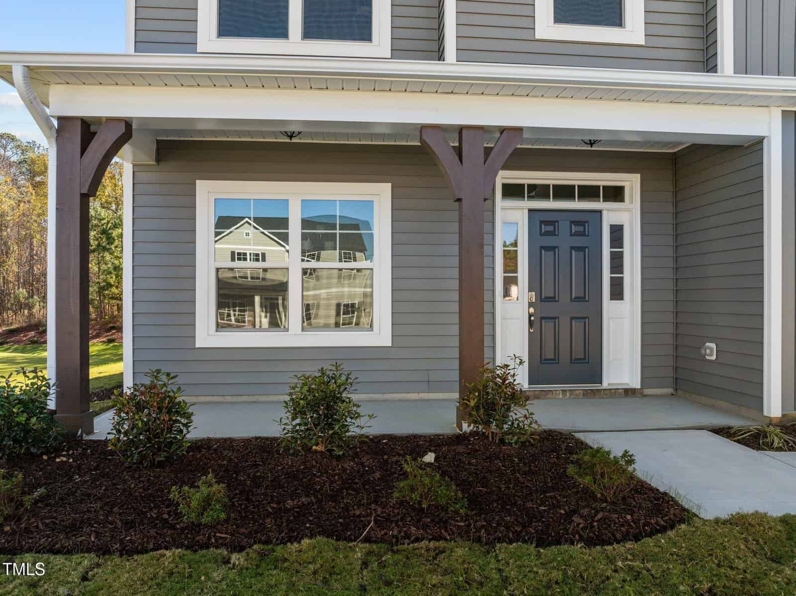 Front view of a modern house with gray siding, a dark gray front door with glass panels, a large window, white trim, a covered porch with wooden columns, and neatly landscaped bushes and mulch.
