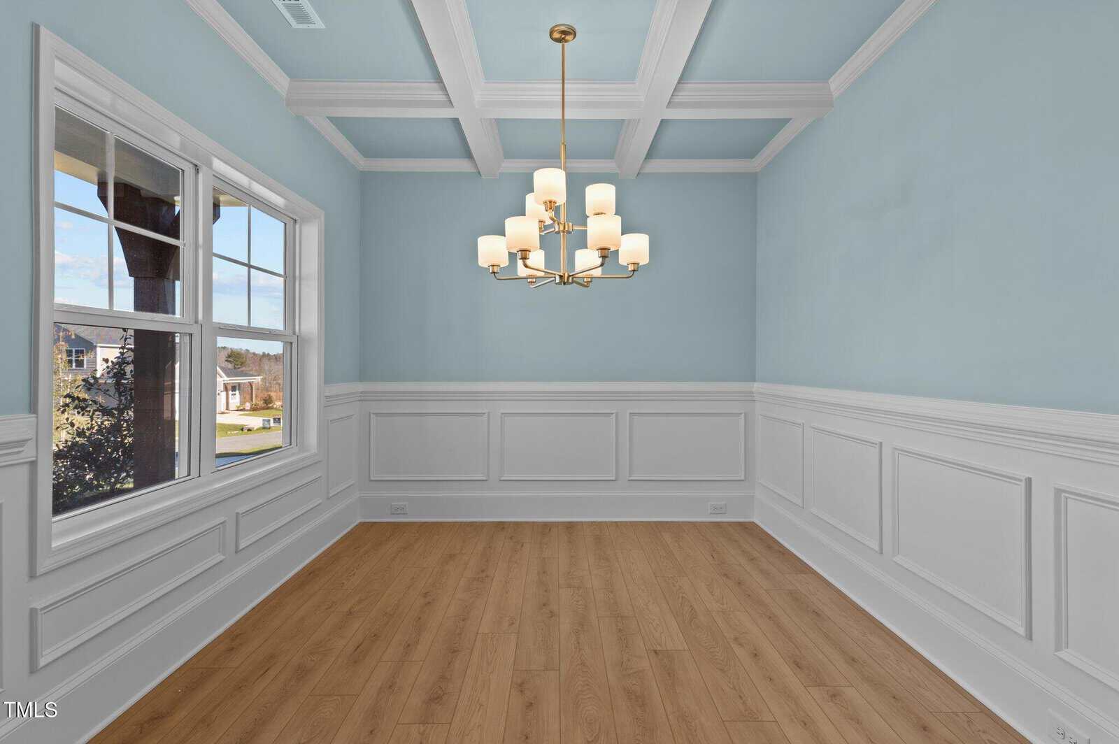 A bright, empty dining room with light blue walls, white wainscoting, wooden floors, a large window, a coffered ceiling, and a modern gold chandelier with multiple lights.