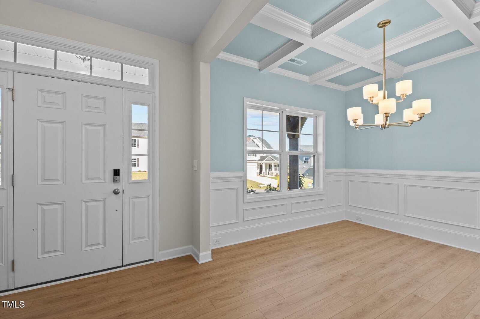 Bright entryway and dining room with light wood floors, white wainscoting, blue walls, coffered ceiling, a modern chandelier, large window, and white front door with glass panels.