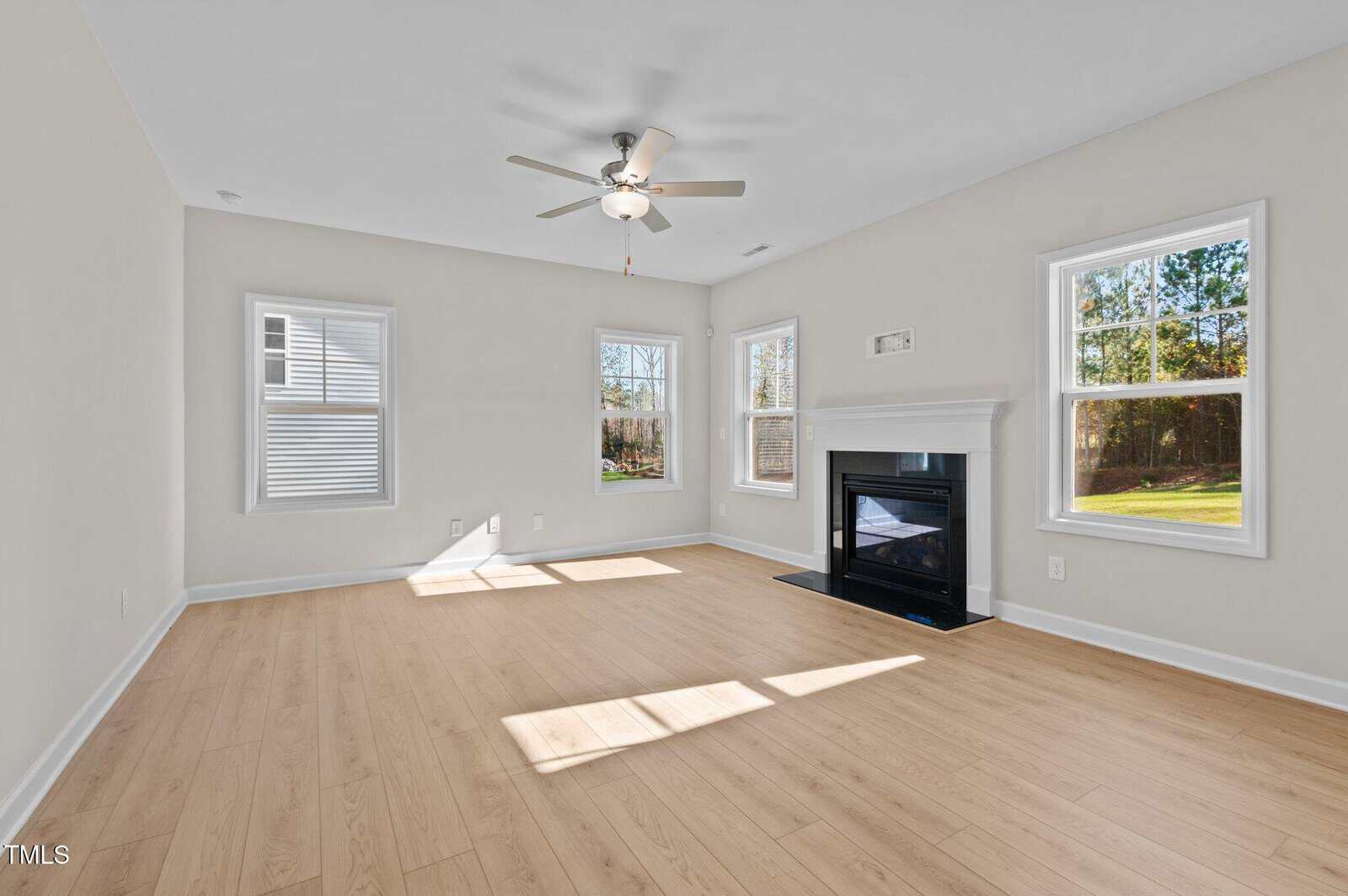 A bright, empty living room with light wood floors, cream walls, a ceiling fan, multiple windows letting in sunlight, and a black framed fireplace.