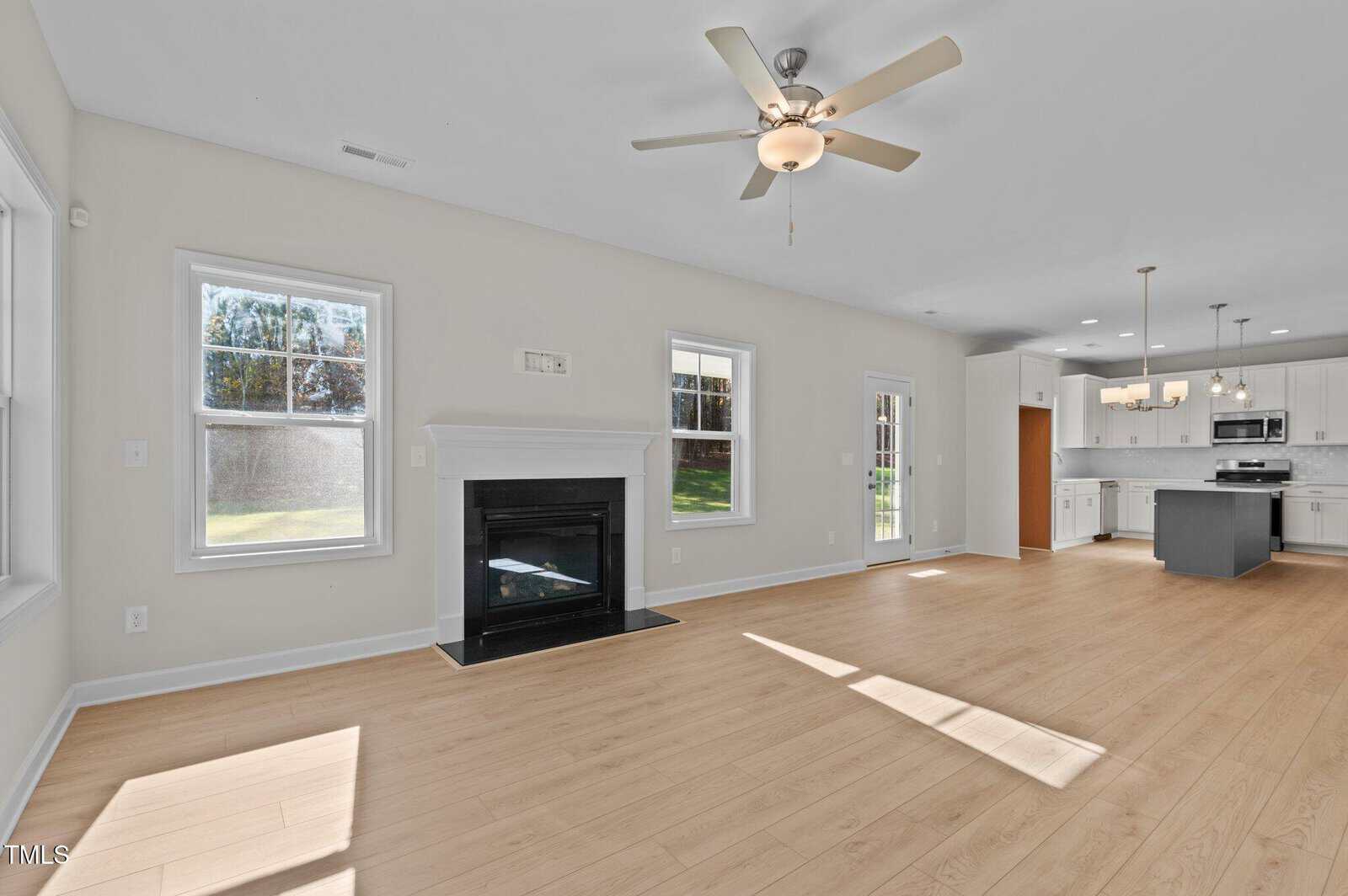 A bright, spacious living room with light wood floors, a ceiling fan, fireplace, and large windows, seamlessly connected to a modern white kitchen with an island and pendant lights.