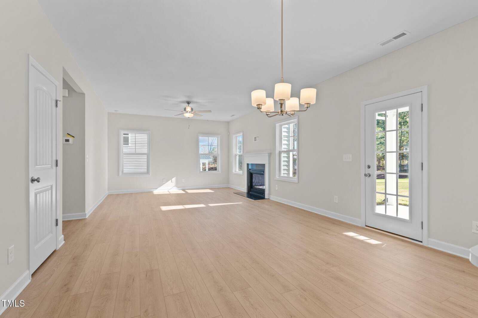 Bright, empty living room with light wood floors, white walls, a ceiling fan, fireplace, and chandelier. Glass doors lead outside, and sunlight streams through multiple windows.