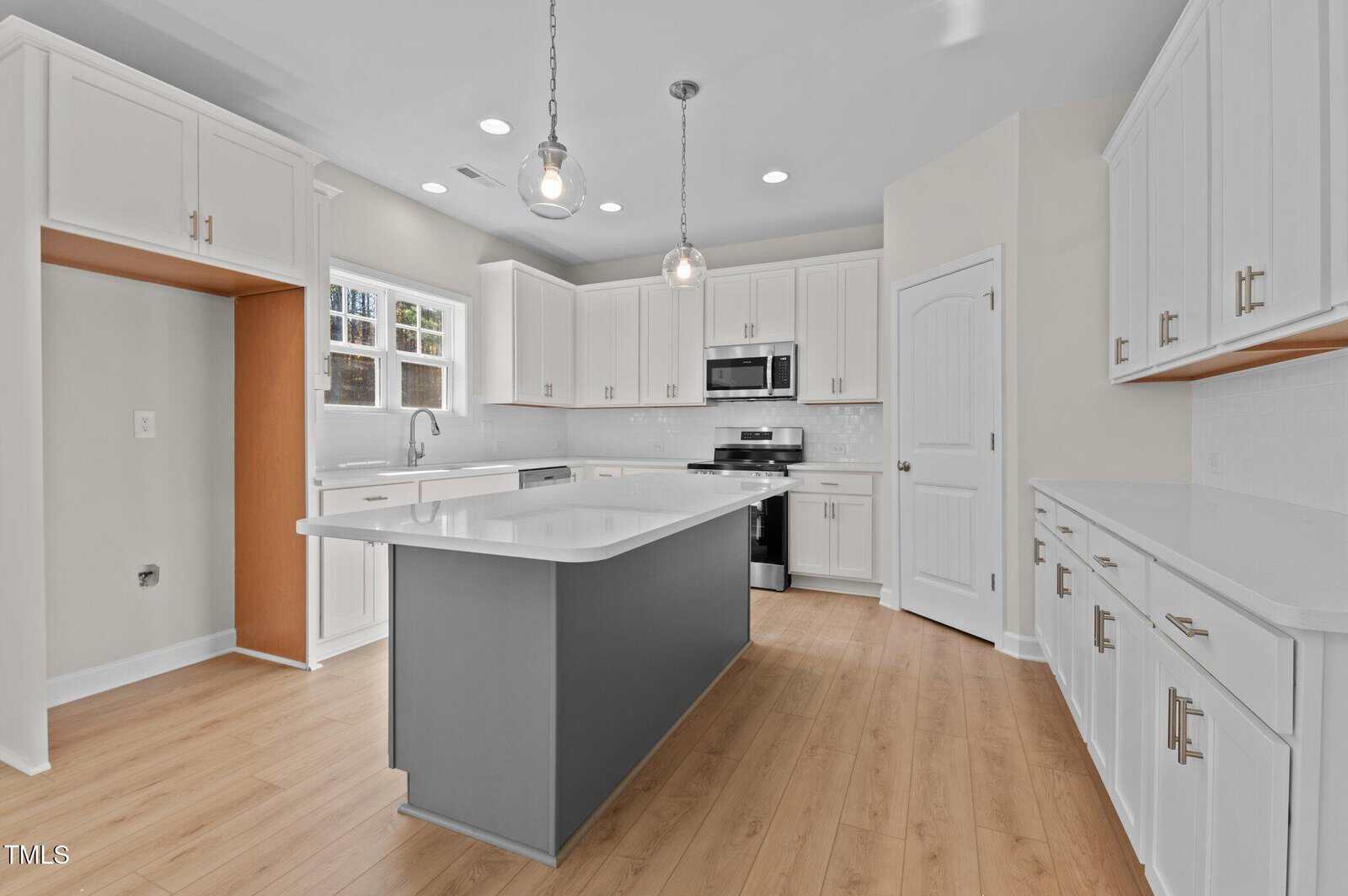 Modern kitchen with white cabinets, stainless steel appliances, a gray island with a white countertop, pendant lights above, wood flooring, and large windows letting in natural light.