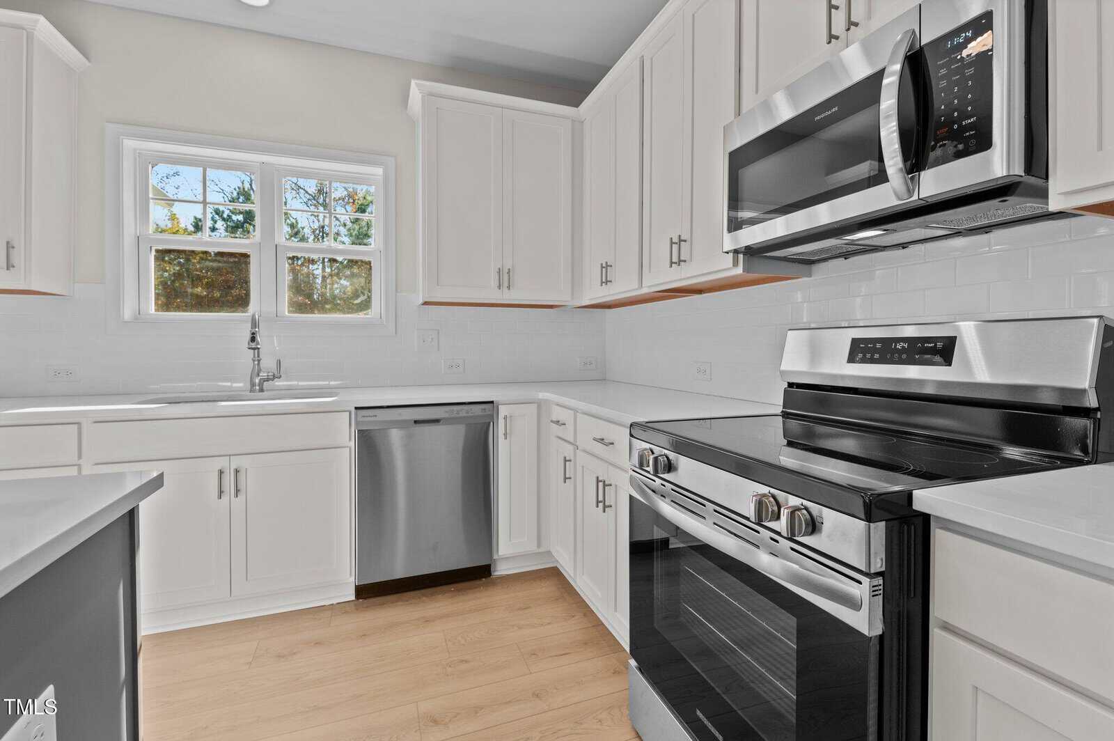 Modern kitchen with white cabinets, stainless steel appliances including an oven, microwave, and dishwasher, white countertops, a double sink under a window, and light wood flooring.