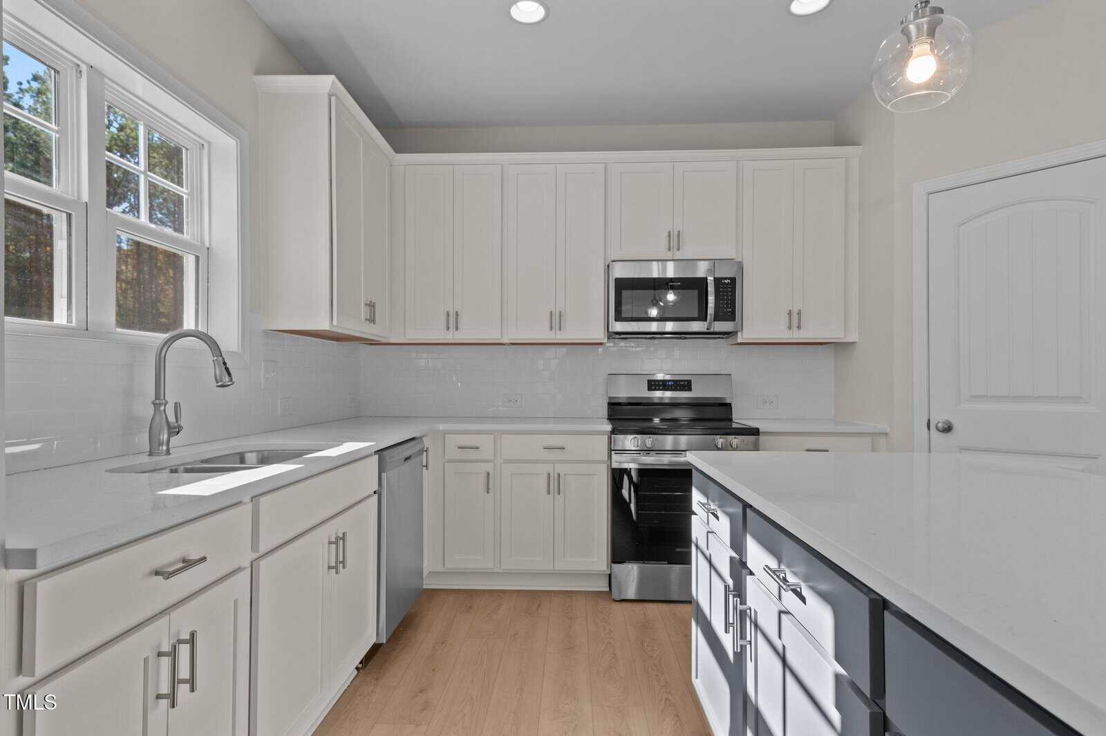Modern kitchen with white cabinets, stainless steel appliances, a large island with dark lower cabinets, quartz countertops, a window over the sink, and light wood flooring. Pendant light hangs above the island.