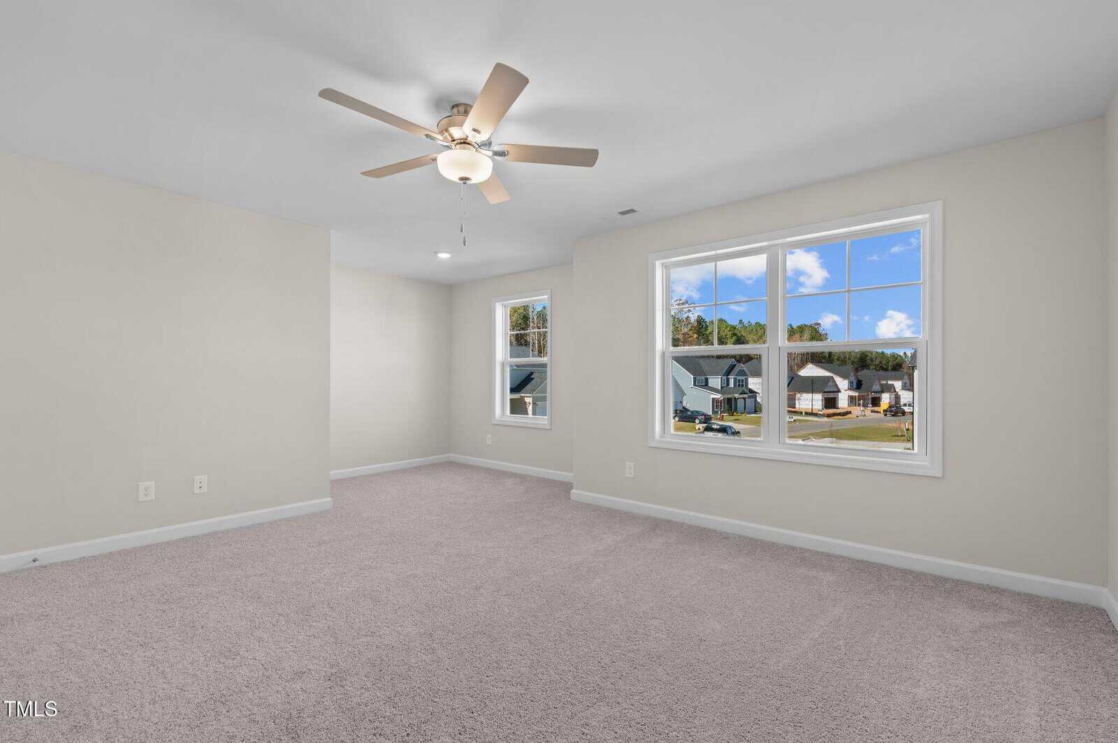 A spacious, empty bedroom with beige carpet, light gray walls, a ceiling fan, and two large windows showing a suburban neighborhood and blue sky.