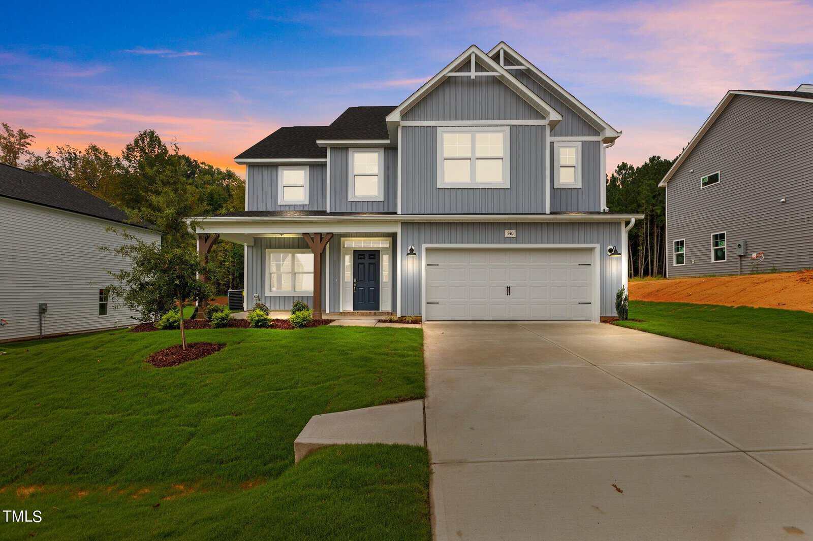 A modern two-story house with blue-gray siding, white trim, a double garage, a covered front porch, and a well-manicured yard. The driveway leads up to the house, and the sky is colorful at sunset.