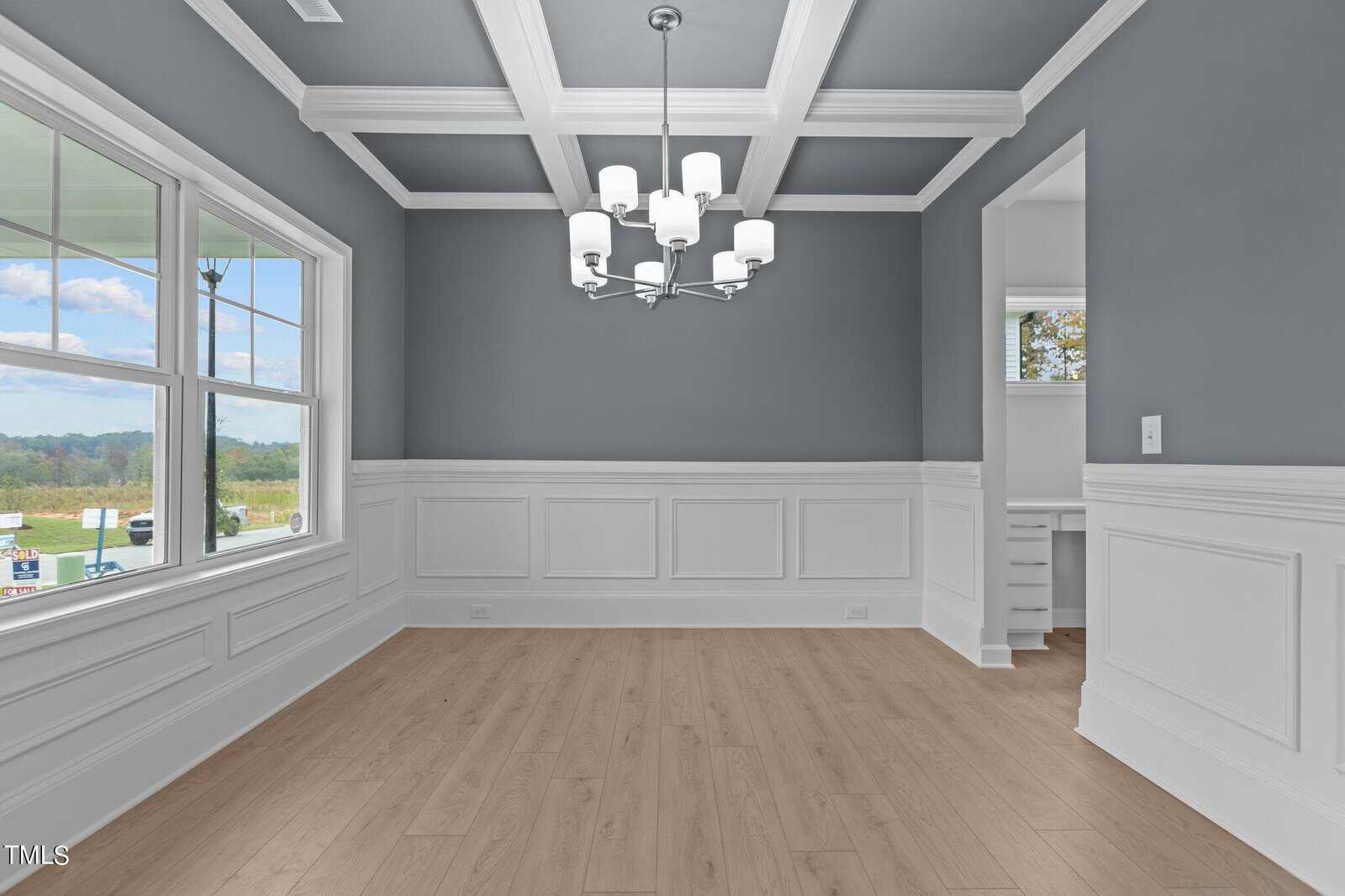 A bright dining room with gray walls, white wainscoting, coffered ceiling, modern chandelier, large window with outdoor view, and light wood flooring. An open doorway leads to an adjacent room.