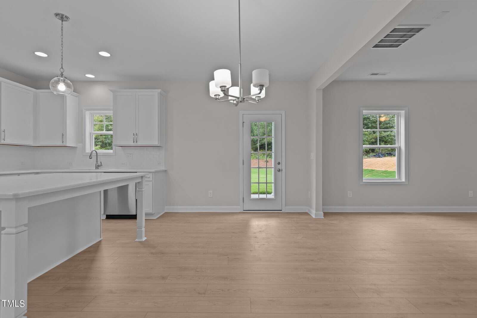 Bright, modern kitchen and dining area with white cabinets, stainless steel appliances, light wood flooring, and large windows. Glass door opens to a green backyard. Contemporary chandelier hangs above dining space.