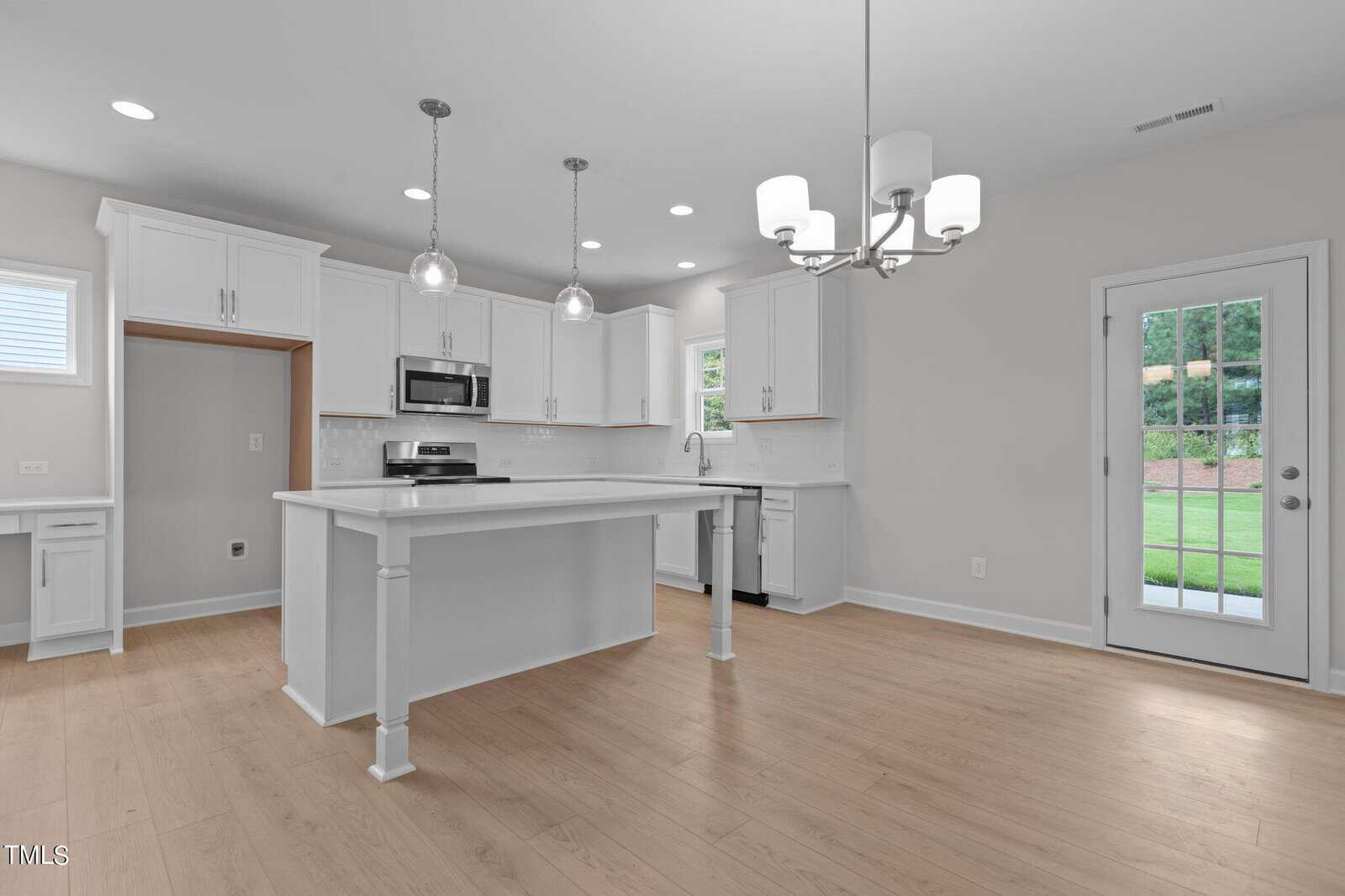 Modern white kitchen with island, stainless steel appliances, pendant lights, light wood flooring, and a glass door leading to a grassy backyard. Room is spacious and bright with natural and overhead lighting.