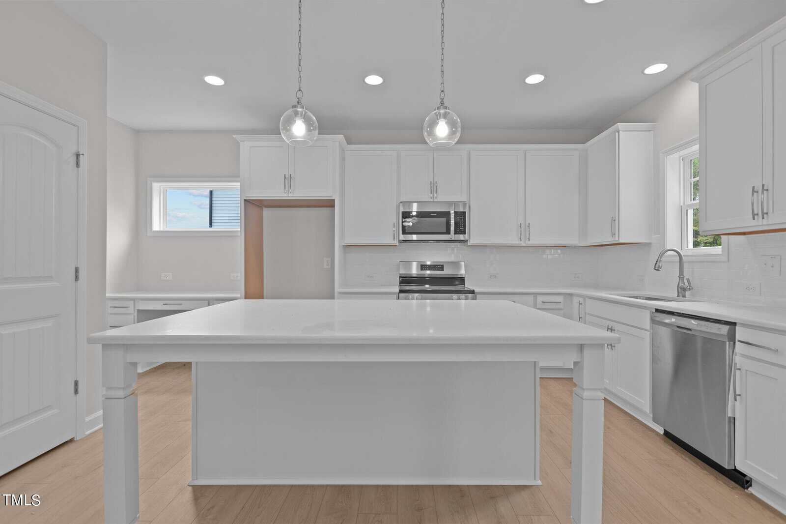 Modern kitchen with white cabinets, stainless steel appliances, a large white island, two pendant lights, light wood floors, and multiple windows letting in natural light.