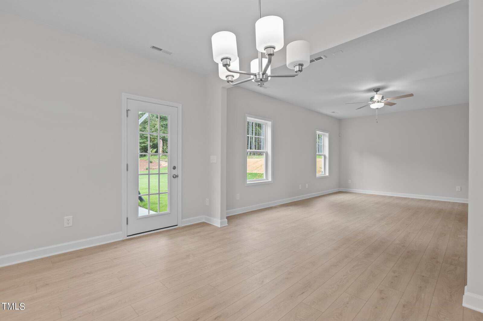 A bright, empty room with light wood flooring, white walls, two windows, a glass door leading outside, a ceiling fan, and a modern white chandelier.
