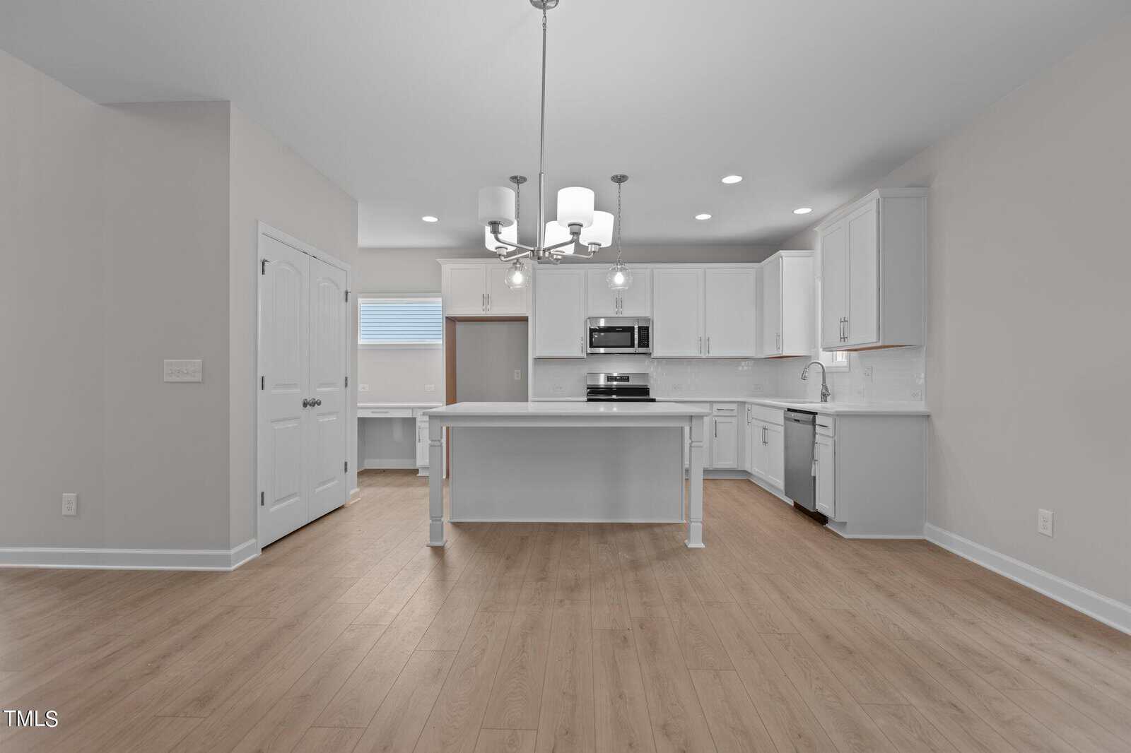 Modern kitchen with white cabinets, stainless steel appliances, a center island with pendant lighting, wood-look flooring, and neutral-colored walls. A double-door pantry and windowed nook are visible on the left.
