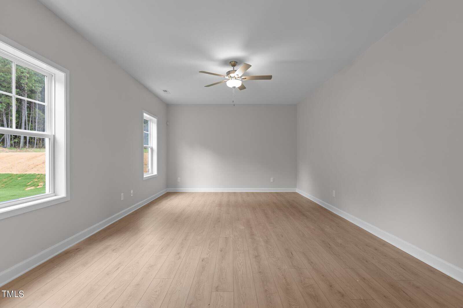 A spacious, empty room with light wood flooring, light gray walls, two large windows, and a ceiling fan with lights. Natural light fills the room.