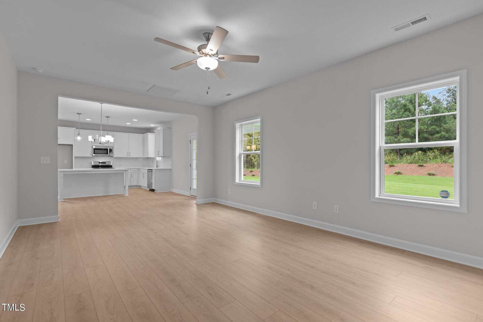 Spacious, empty living room with light wood flooring, white walls, ceiling fan, and two large windows. In the background, a bright kitchen with white cabinets and appliances is visible.