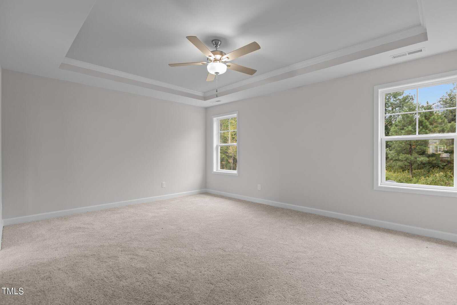 Empty bedroom with beige carpet, light gray walls, tray ceiling, ceiling fan with lights, and two large windows letting in natural light. Trees are visible outside the windows.