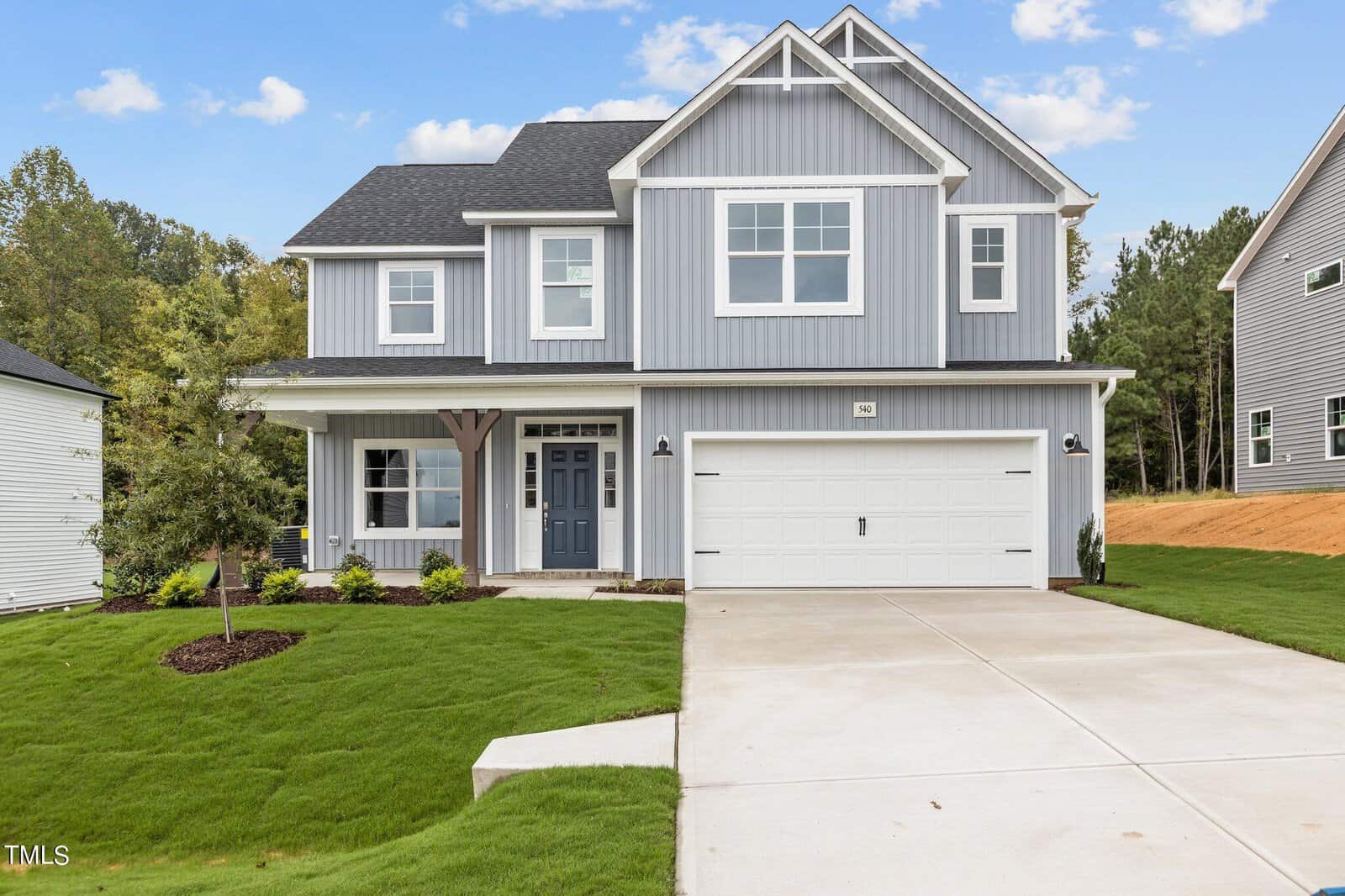 Two-story modern suburban house with gray siding, white trim, a covered porch, double garage, and a neatly manicured lawn, set against a backdrop of trees and a clear sky.