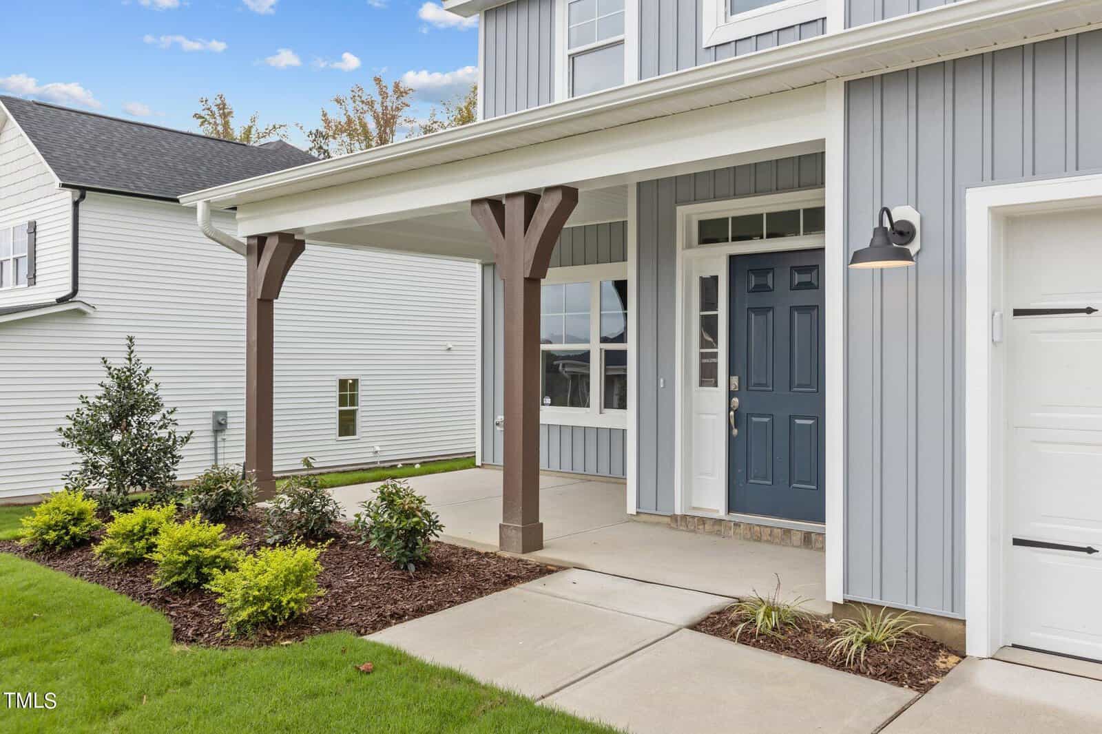 Modern house exterior with blue and white siding, a covered front porch with wooden columns, landscaped garden beds, and a dark blue front door next to a garage door.