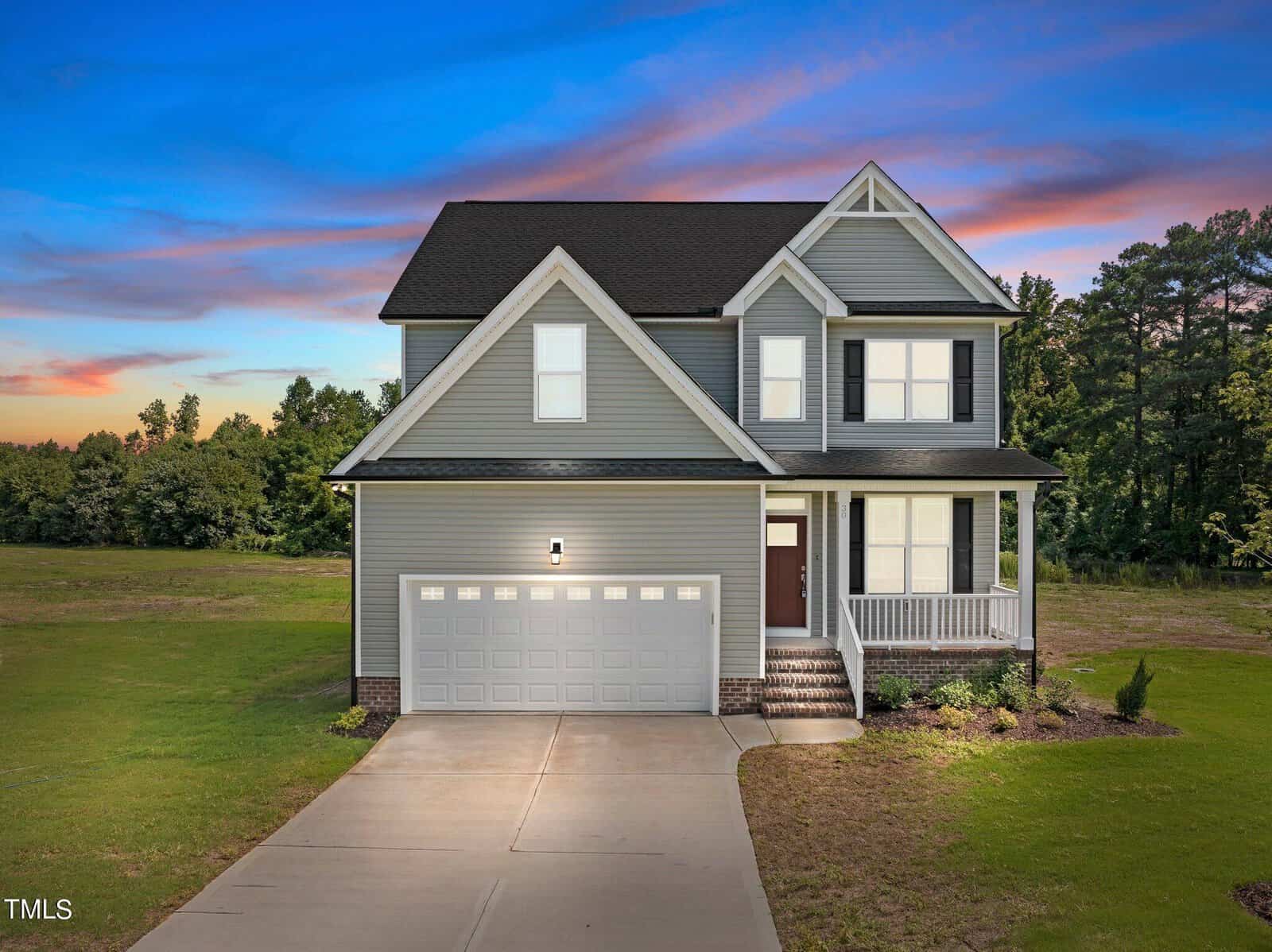 A two-story gray house with white trim, a front porch, and a two-car garage sits on a large grass lawn. Trees line the background under a colorful sunset sky.