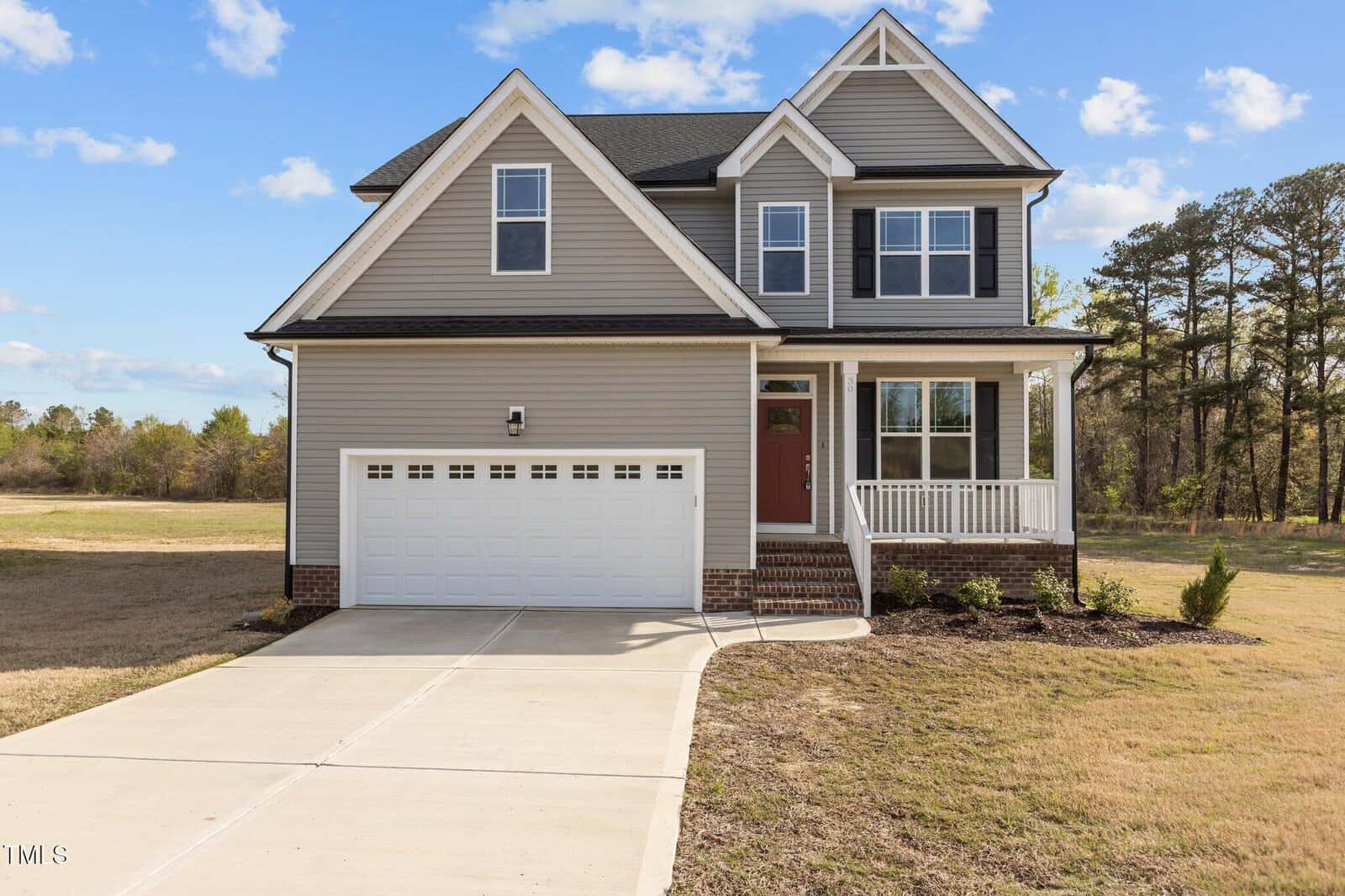 Two-story suburban house with gray siding, white trim, a front porch, and a red front door. The home features a double garage and a wide concrete driveway, with a grassy yard and trees in the background under a blue sky.