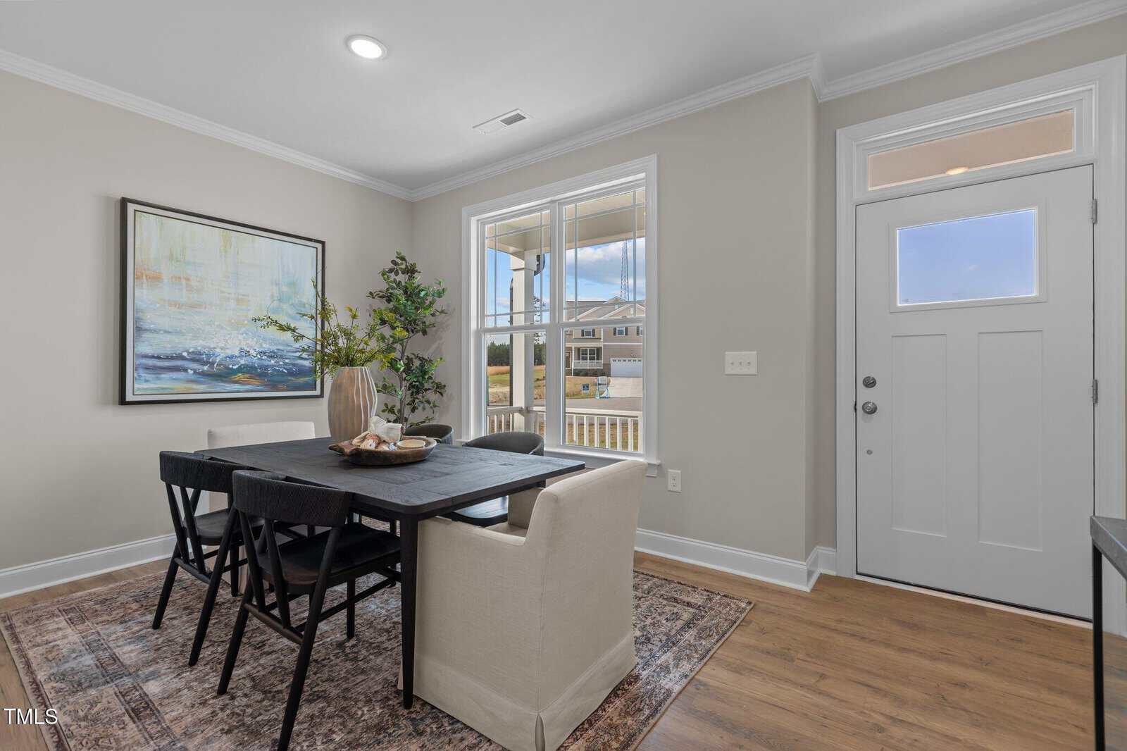 Modern dining area with a black table, four black chairs, and a white armchair, set on a patterned rug. Large window and glass door let in natural light. A painting and potted plant decorate the light beige walls.