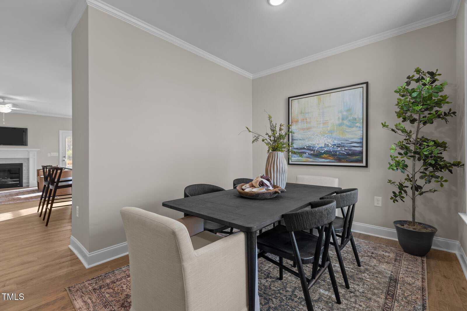 A modern dining room with a black table, six chairs, a large painting on the wall, a decorative vase, and a potted plant in the corner. The room has light walls, wood flooring, and a view into the living area.