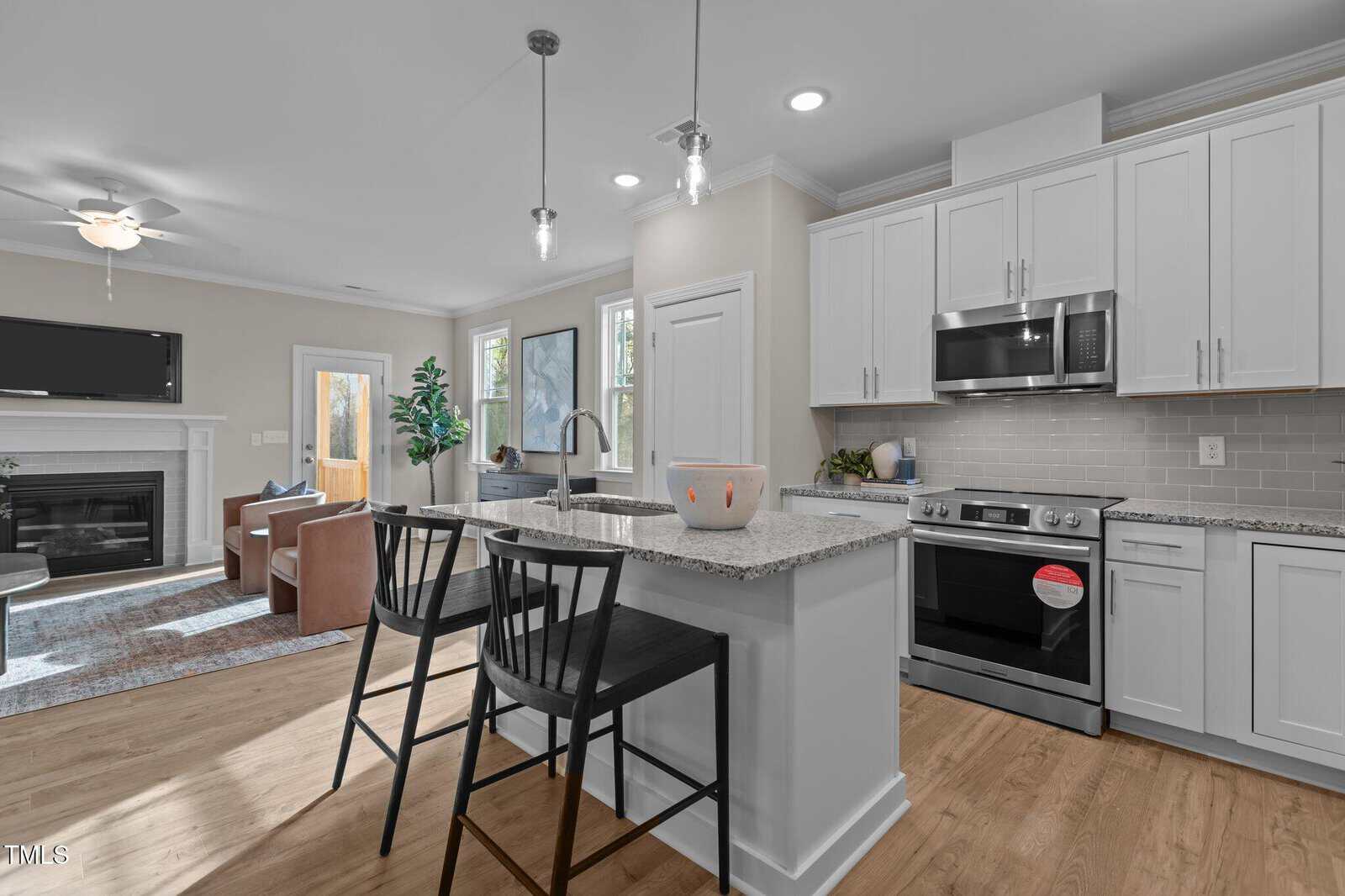 Modern kitchen with white cabinets, stainless steel appliances, granite countertops, and an island with two black chairs; adjacent open living area with leather chairs, a rug, and a fireplace.