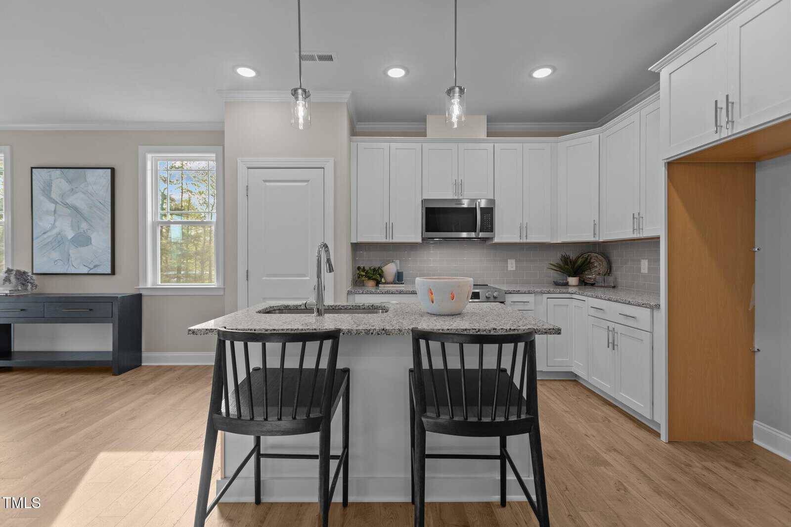 Modern kitchen with white cabinets, stainless steel appliances, a granite island with a sink, two black chairs, and pendant lights. A bowl sits on the island; a desk and artwork are visible by the windows.