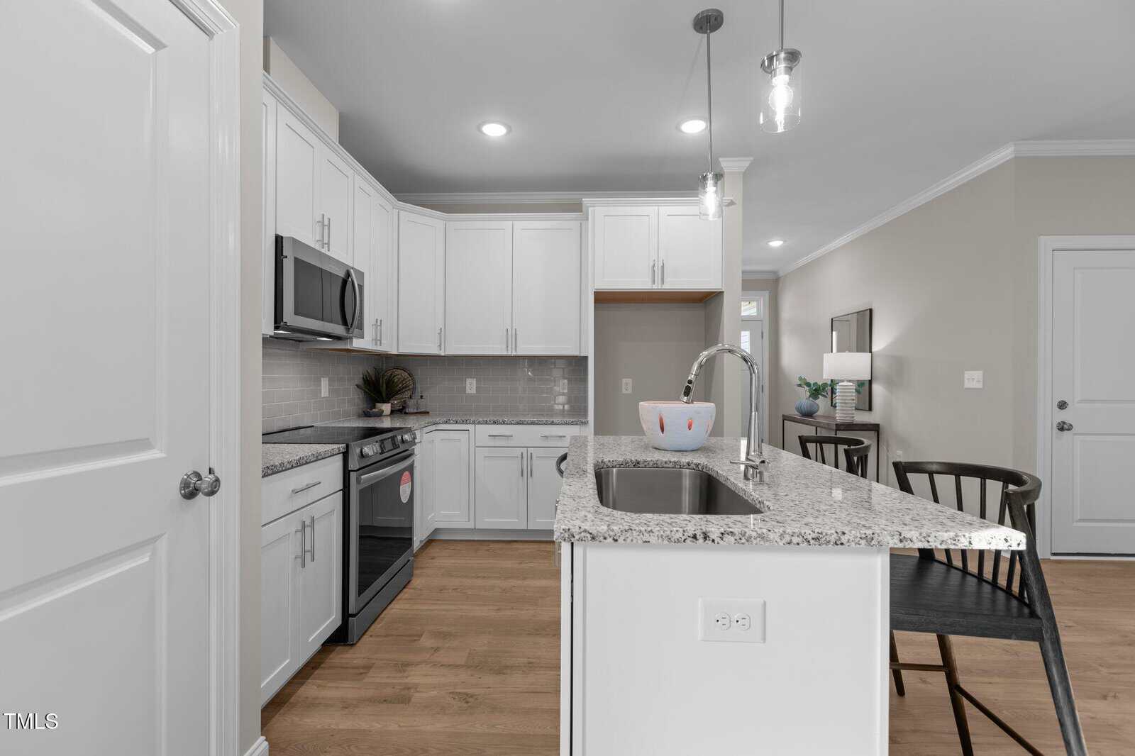 Modern kitchen with white cabinets, stainless steel appliances, a granite island countertop with a sink, pendant lights overhead, and two black chairs. Light wood flooring and neutral walls create a bright, clean look.