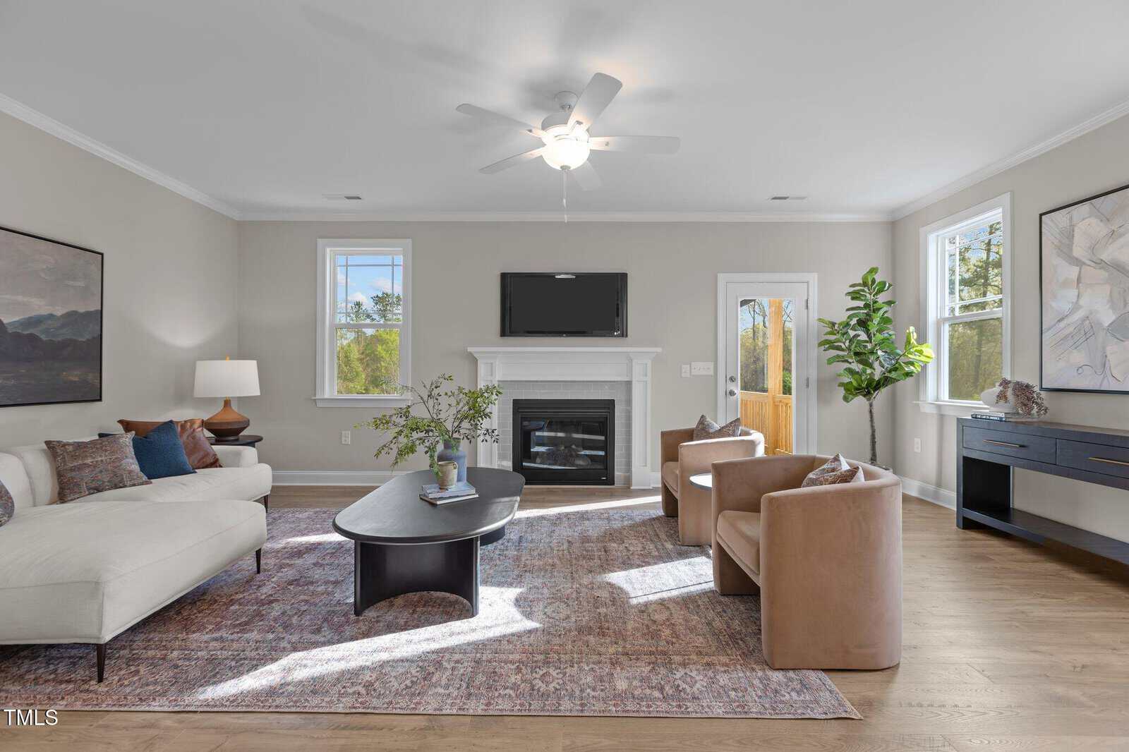 A bright living room with a white sofa, two tan armchairs, a black coffee table, a fireplace with a TV above it, large windows, a plant, and modern decor on wood floors and a patterned rug.