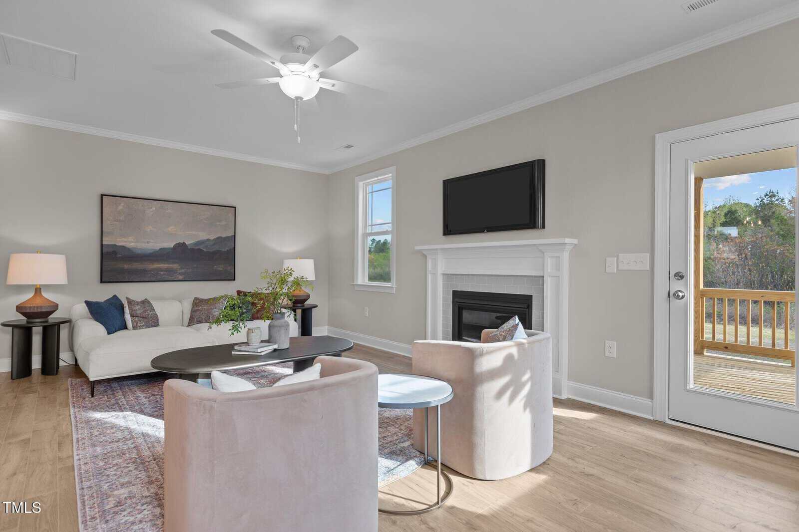 Bright living room with a white sofa, two light chairs, a coffee table, a fireplace with a TV above, large windows, and a glass door leading to a wooden deck. The space is decorated in neutral tones.