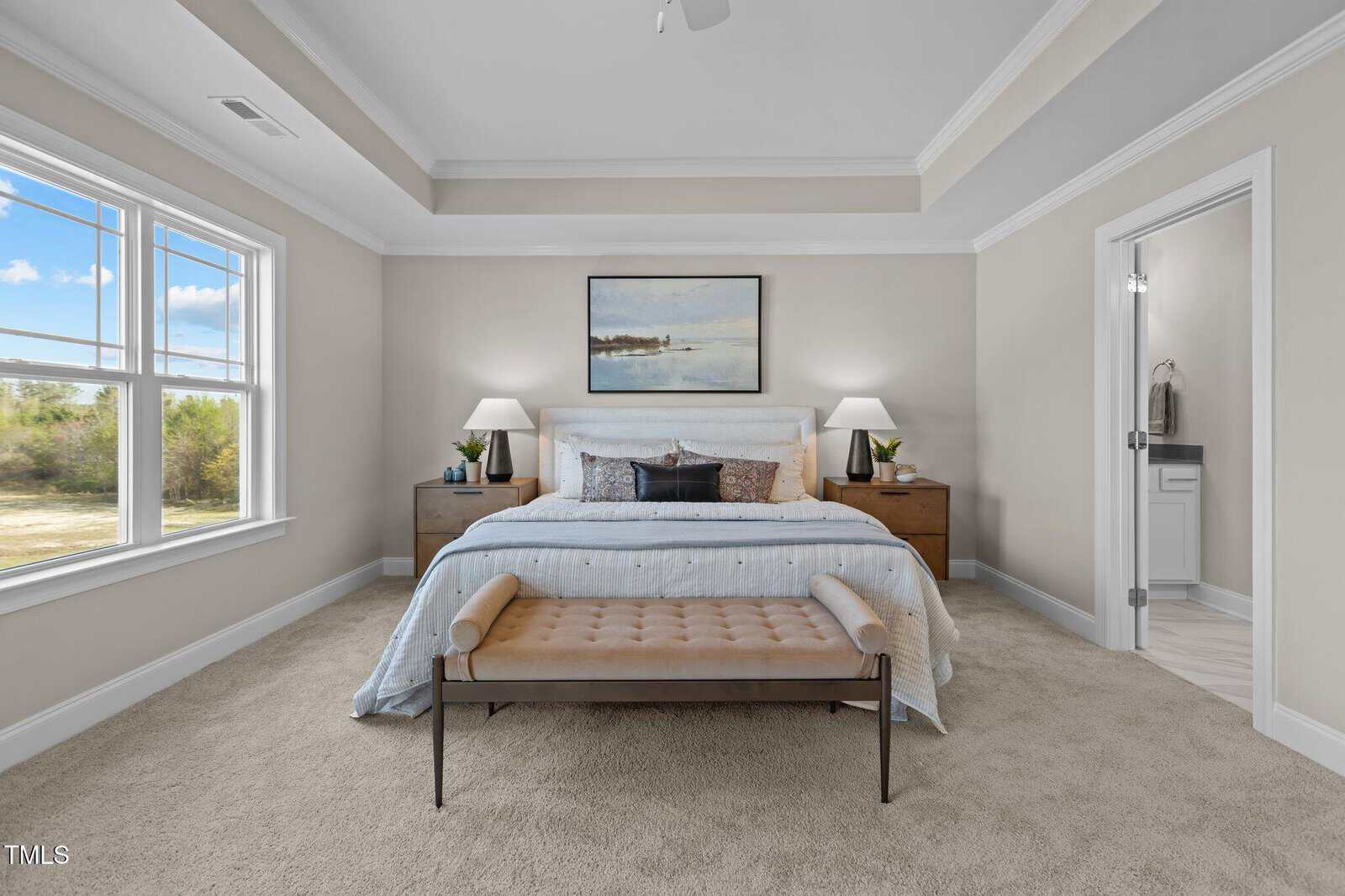 A bright bedroom with a large bed, two nightstands with lamps, a bench at the foot of the bed, a landscape painting above the headboard, and an ensuite bathroom visible to the right.