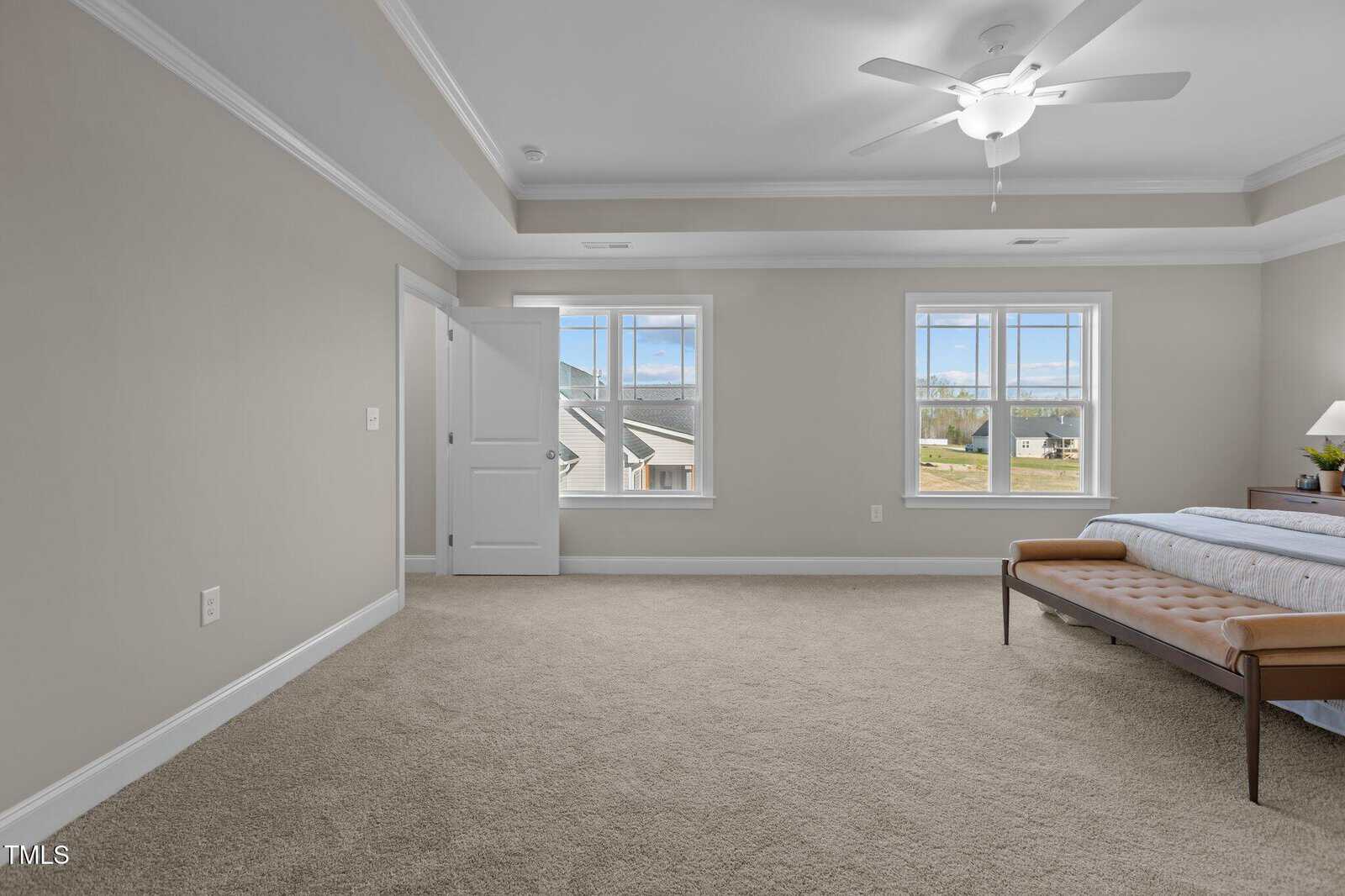 Spacious bedroom with beige carpet, light gray walls, two large windows, a ceiling fan, a bed with white linens, and a brown bench at the foot of the bed. Doorways lead to other rooms.