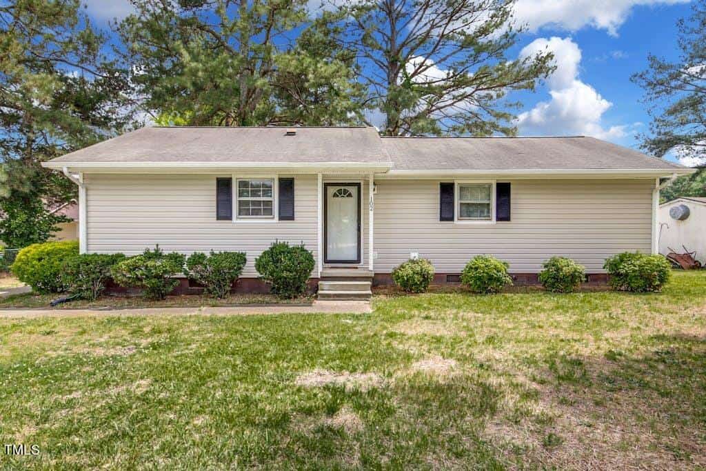 A single-story beige house at 102 Briarcliff Street, Henderson NC, with white trim, a central white door, and symmetrical windows, surrounded by a green lawn and small shrubs under a clear sky.