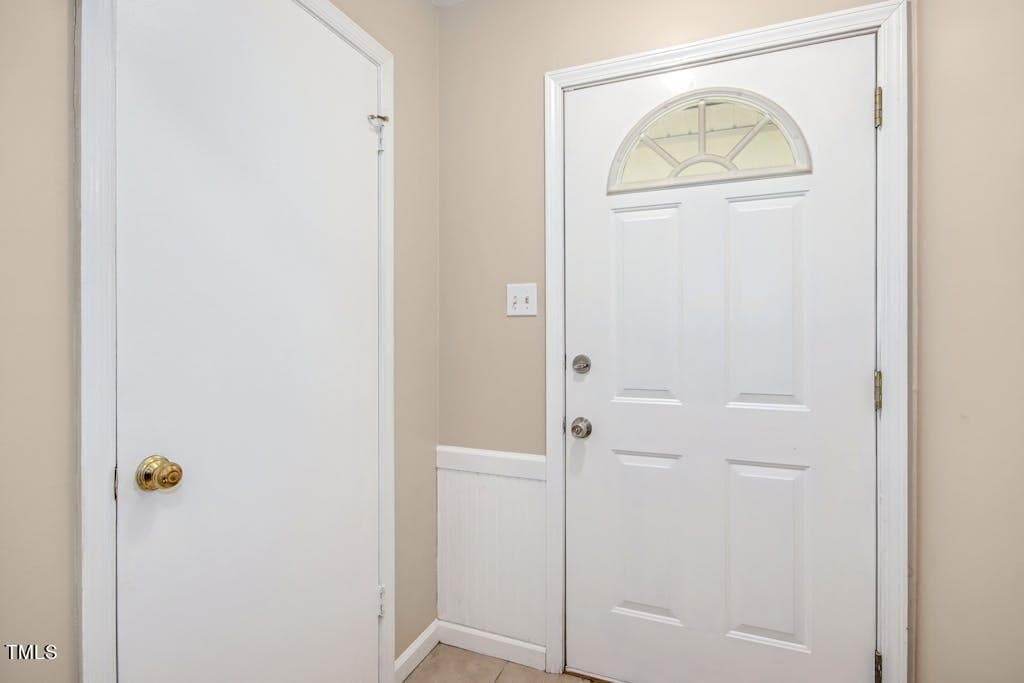 A home interior featuring a white entry door with an arched window and brass handle, flanked by a plain white wall and a closed white door to the left.