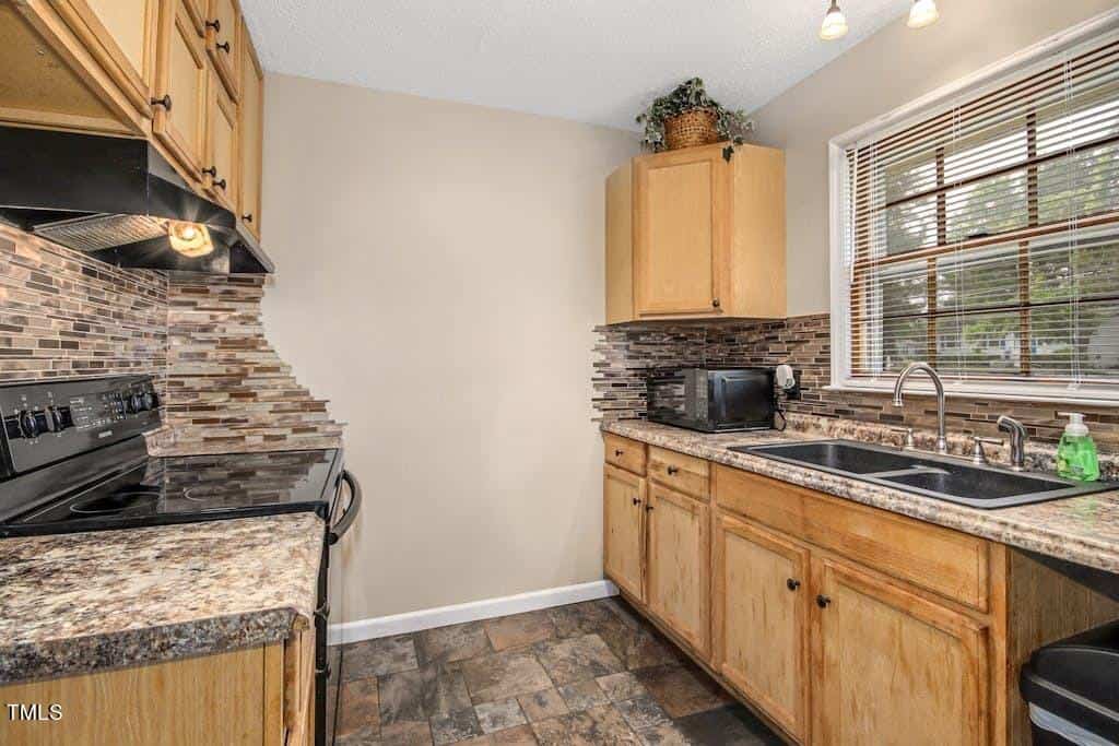 Modern kitchen with wooden cabinets, granite countertops, and a mosaic backsplash. features include a sink under a window overlooking greenery, and a tiled floor.