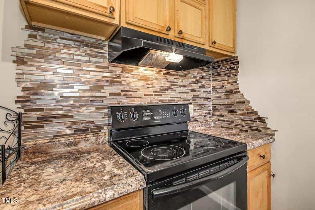 A modern kitchen with granite countertops, wooden cabinets, and a black electric stove. a unique multicolored glass tile backsplash adds a decorative touch.